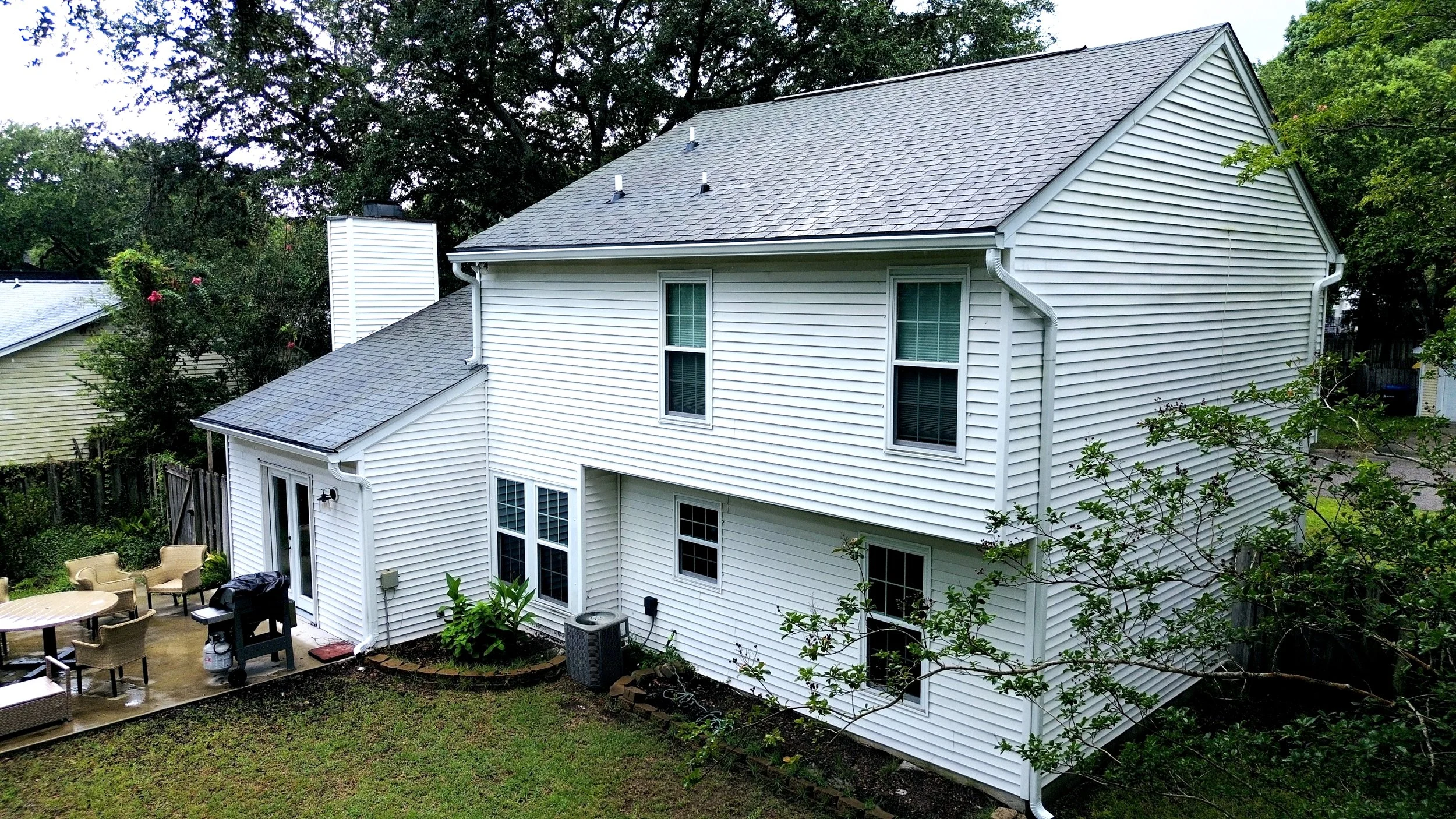 Backyard view of a two-story white house with vinyl siding, gray shingle roof, multiple windows, a sliding glass door, patio furniture, grill, and surrounding green trees and grass.