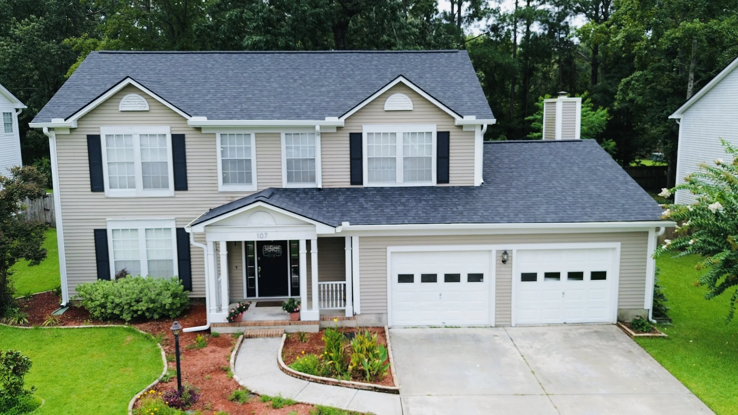 A two-story beige house with white trim and black shutters, featuring a two-car garage and a covered front porch with white columns. The front yard has a green lawn, flower beds, and a concrete walkway leading to the front door, with trees in the bac