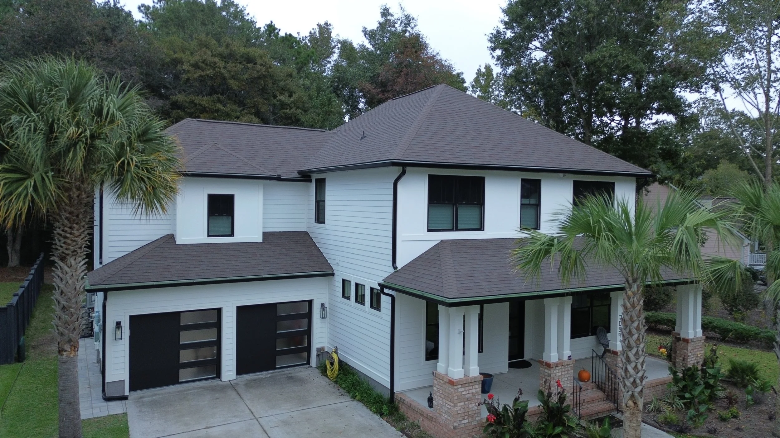 A modern two-story house with white siding, black window frames, and a gray shingle roof, surrounded by palm trees and a well-kept lawn.