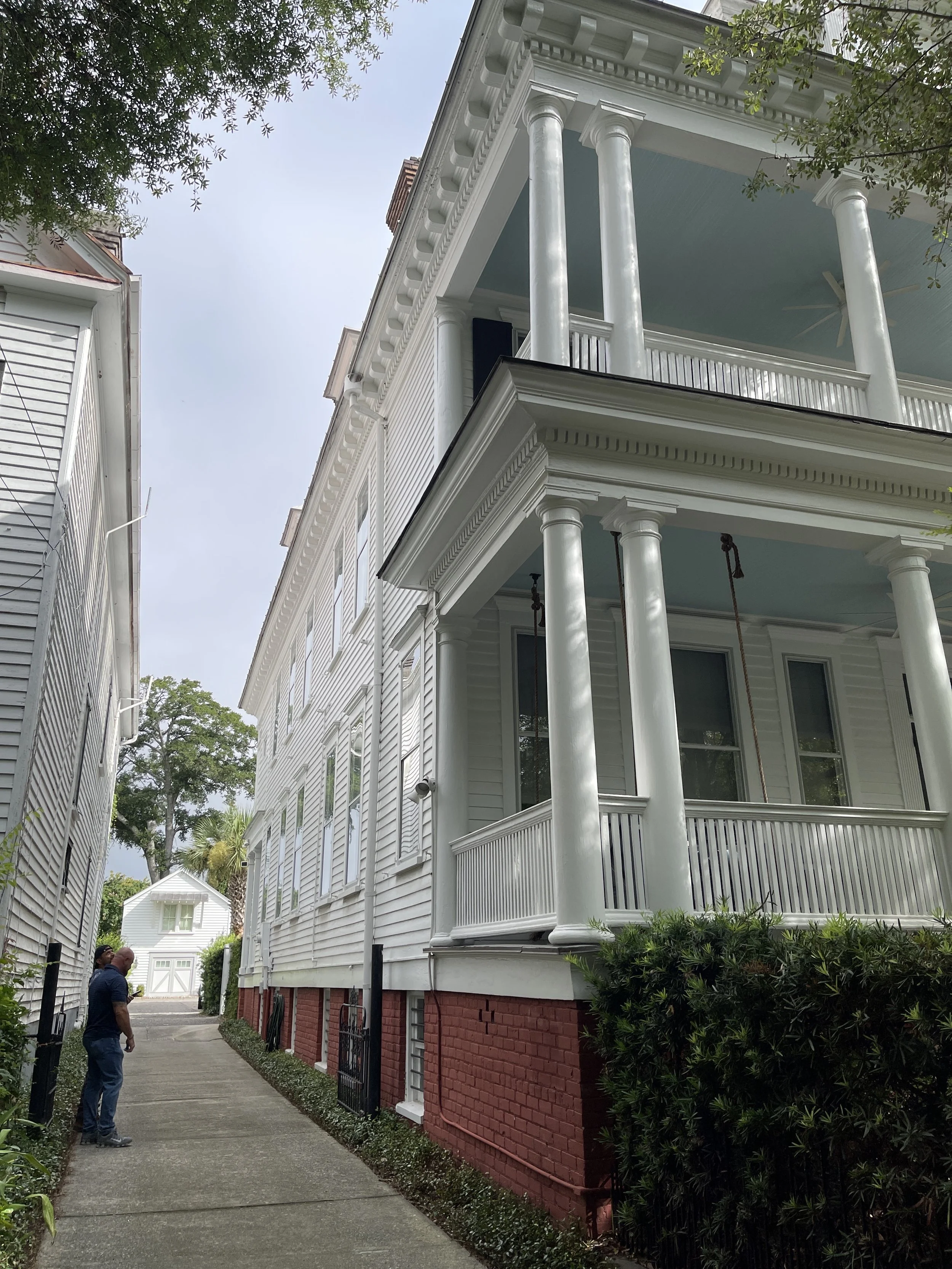 A white, three-story mansion with classical architectural features, including tall columns and intricate cornices, situated along a narrow alley with two people standing nearby.