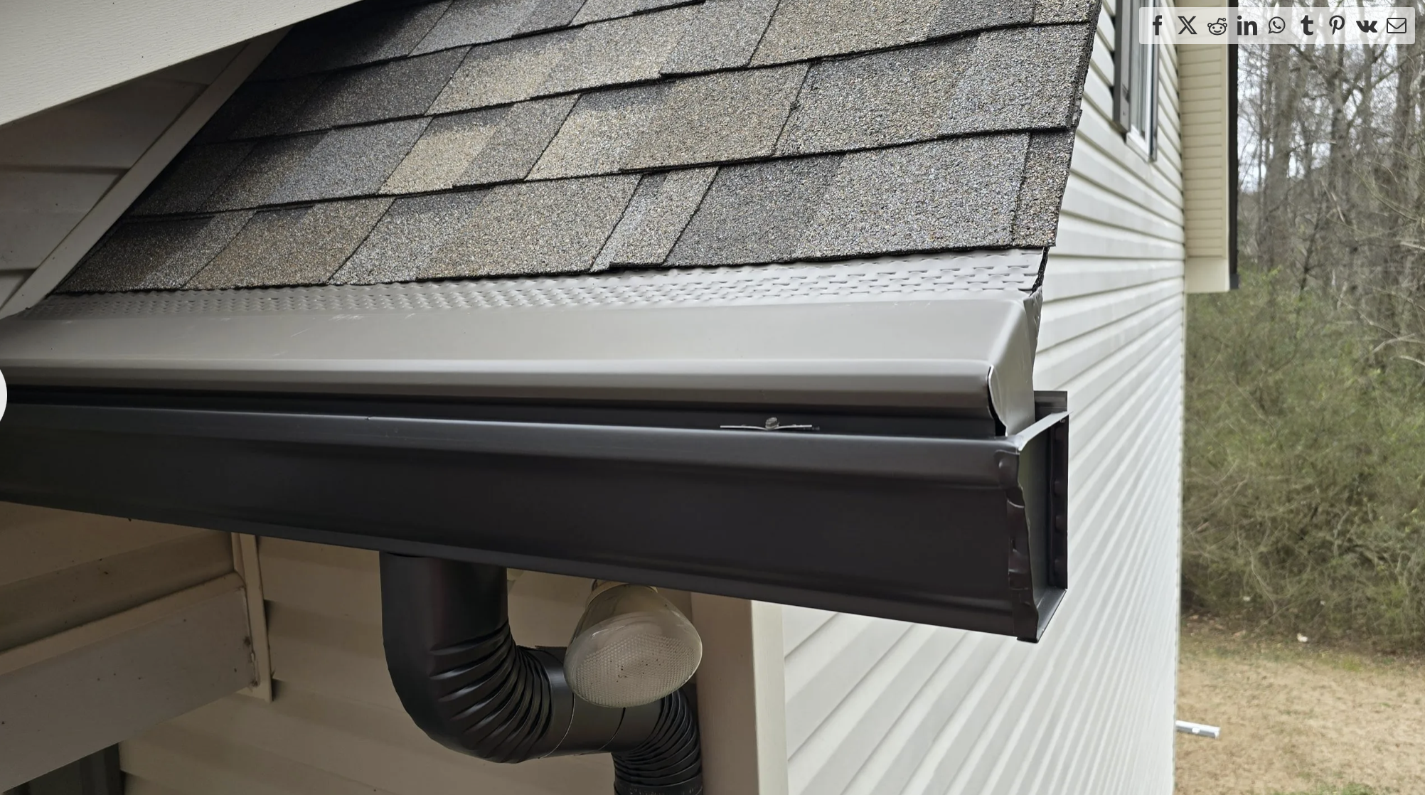 Close-up of a house's roof with brown shingles, black gutter system, and a black downspout attached to the beige vinyl siding.