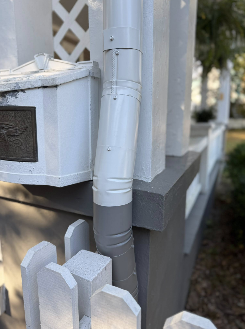 A gray downspout attached to a white house exterior, with a white gate and a gray concrete foundation, and a decorative metal plaque visible in the image.