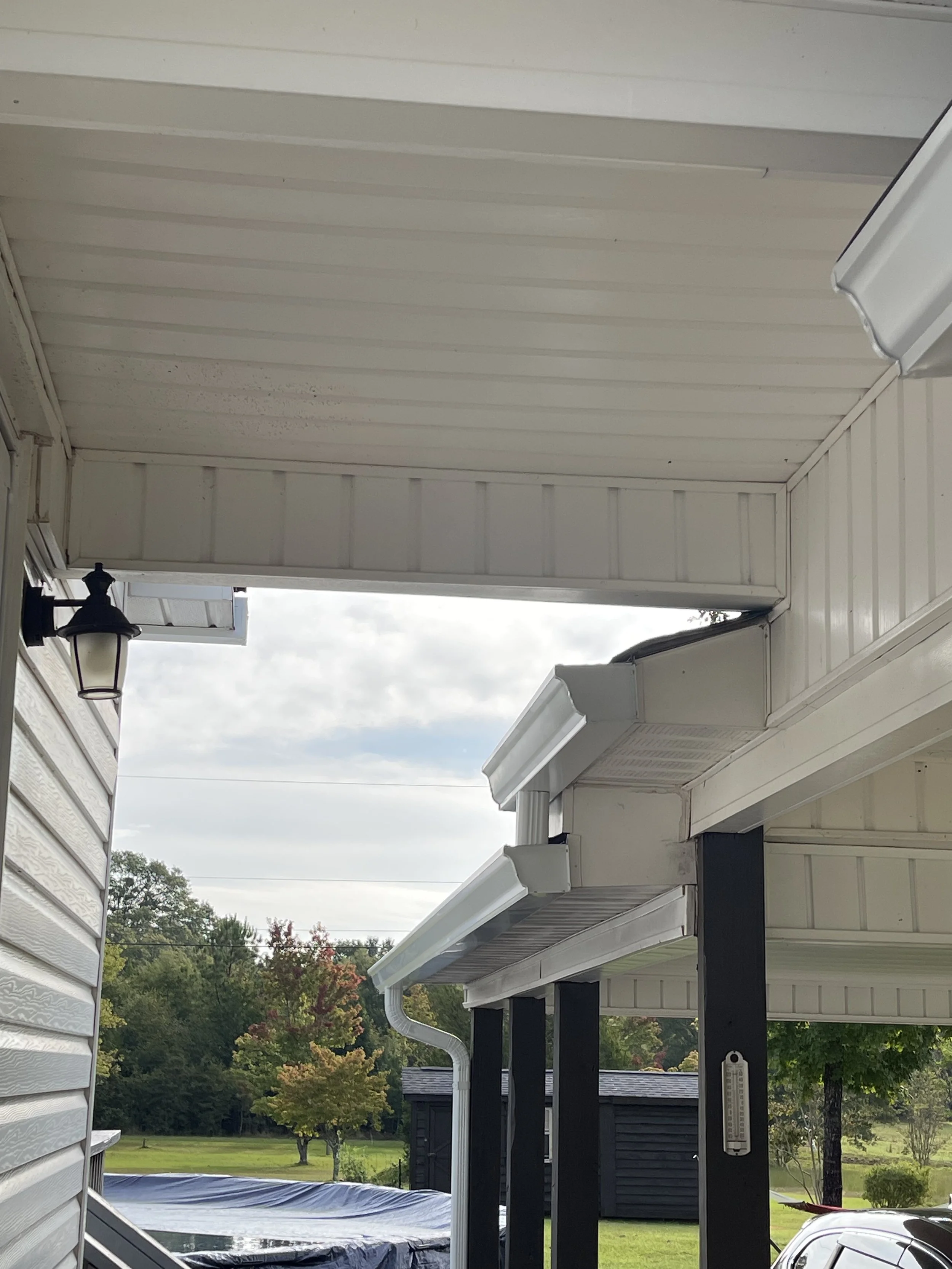 Close-up of house's covered porch with white siding, black posts, attached gutter system, outdoor light fixture, and view of green yard with trees.