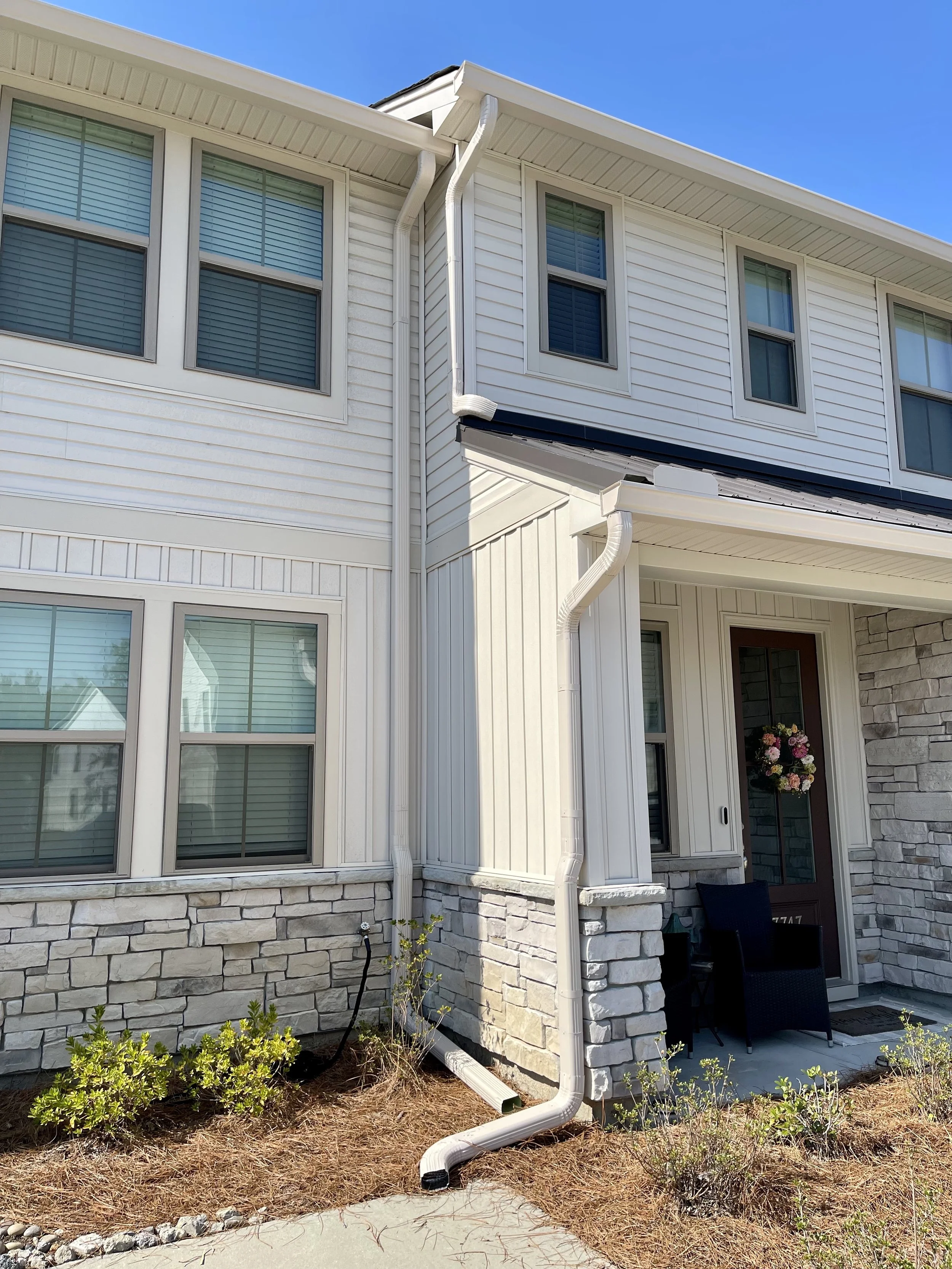 Exterior of a white multi-story house with windows, a stone foundation, a porch with a chair, and a flower wreath on the front door, under a clear blue sky.