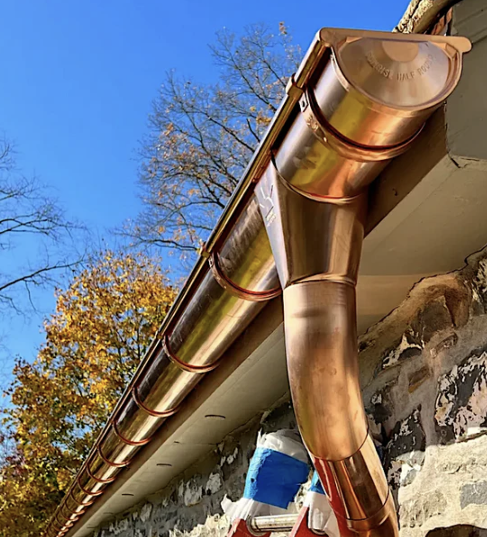 Copper rain gutter with a downspout attached, installed along the edge of a roof. The background shows trees with autumn leaves and a clear blue sky.