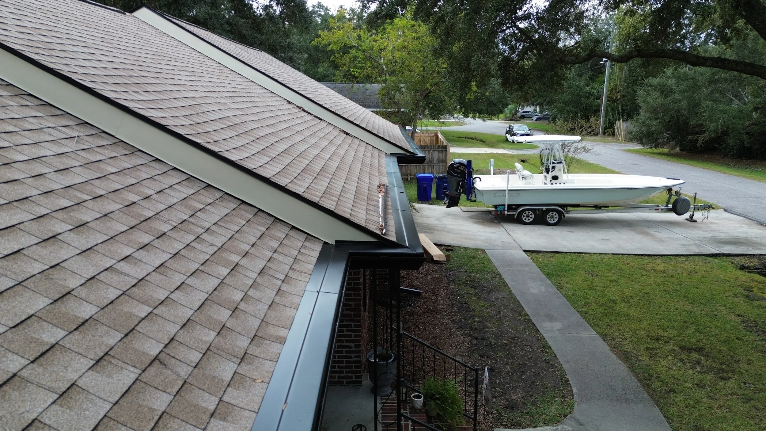 View of a residential roof with tan shingles and a boat on a trailer parked on a driveway, with trees and other houses in the background.