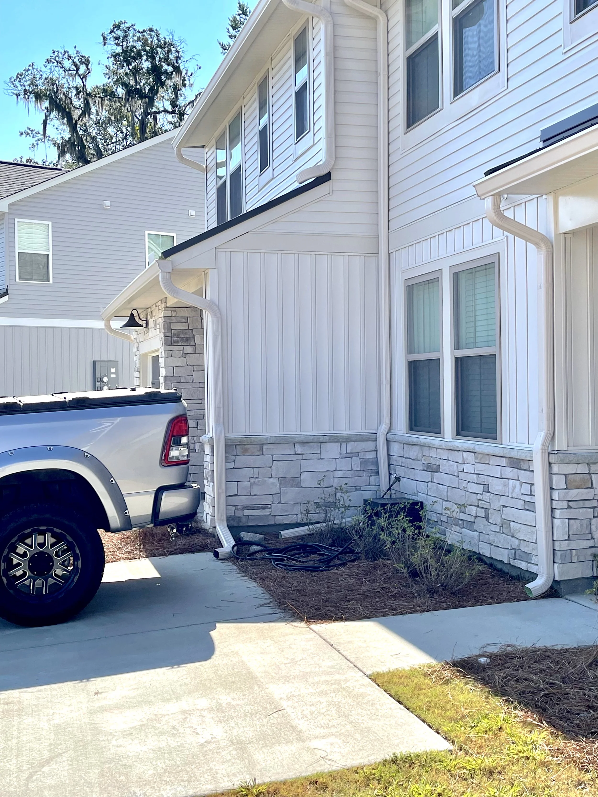 Side view of a white two-story house with stone accents, several windows, and downspouts, with a gray pickup truck parked on the driveway and some plants and hoses near the house.