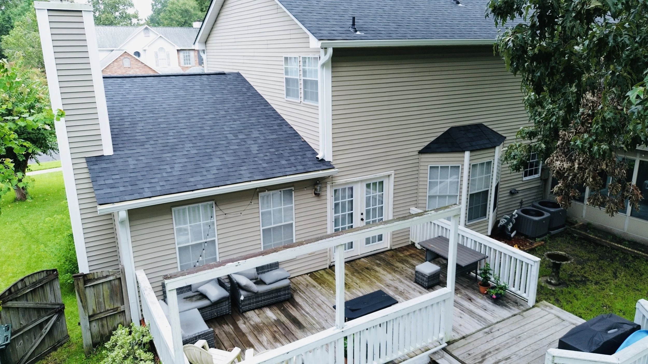 A backyard deck attached to a beige house with beige siding. The deck has outdoor furniture including a patio sofa with cushions, a table, and a small potted plant. The deck is surrounded by a white wooden railing and has some moss or dirt on the woo