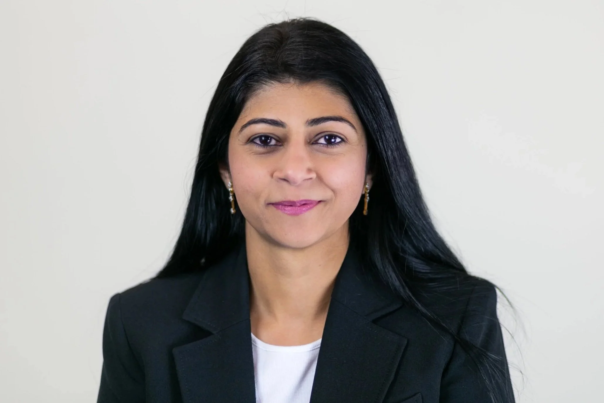 Headshot of a woman with long black hair, wearing a black blazer and earrings, standing against a plain background.
