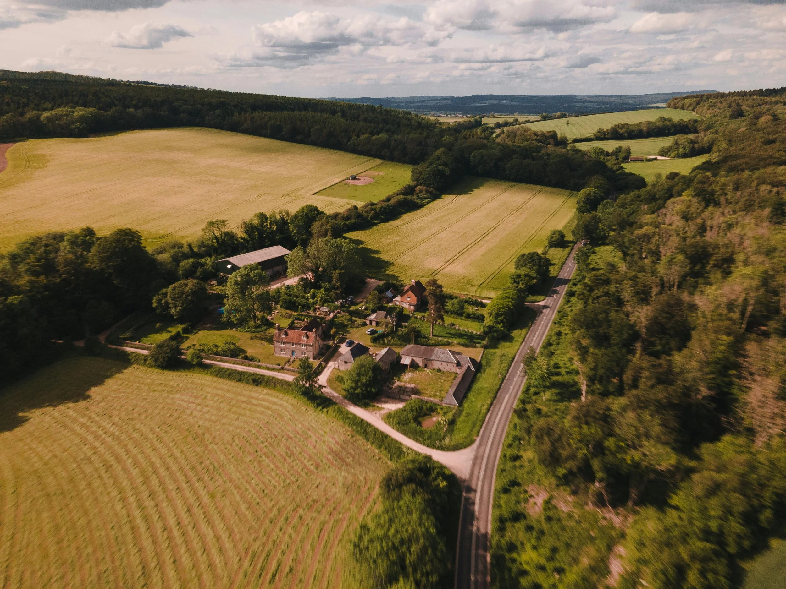 Aerial view of a rural landscape featuring a small cluster of houses surrounded by green fields, trees, and a winding road, with rolling hills in the background under a partly cloudy sky.