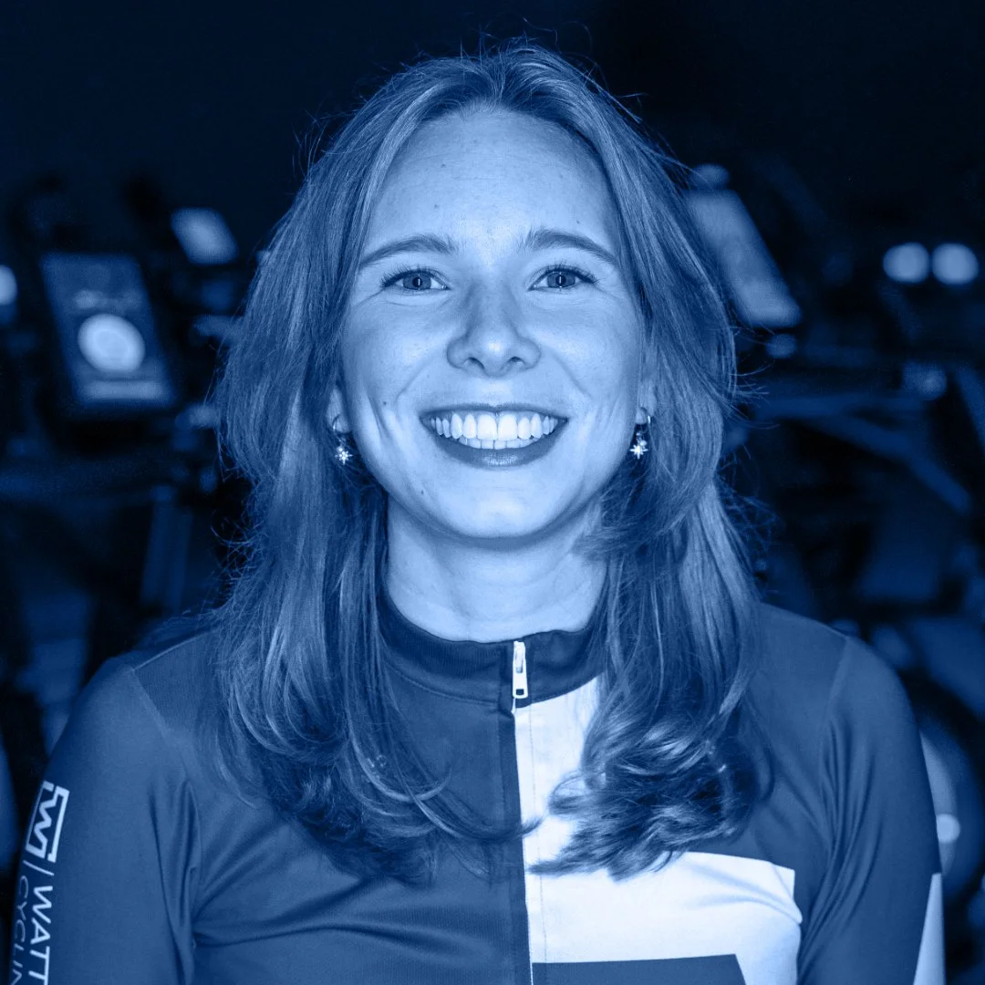 Een vrouw met lang, lichtbruin haar, lachend, in een sportoutfit, met een donkere achtergrond met fietsen.