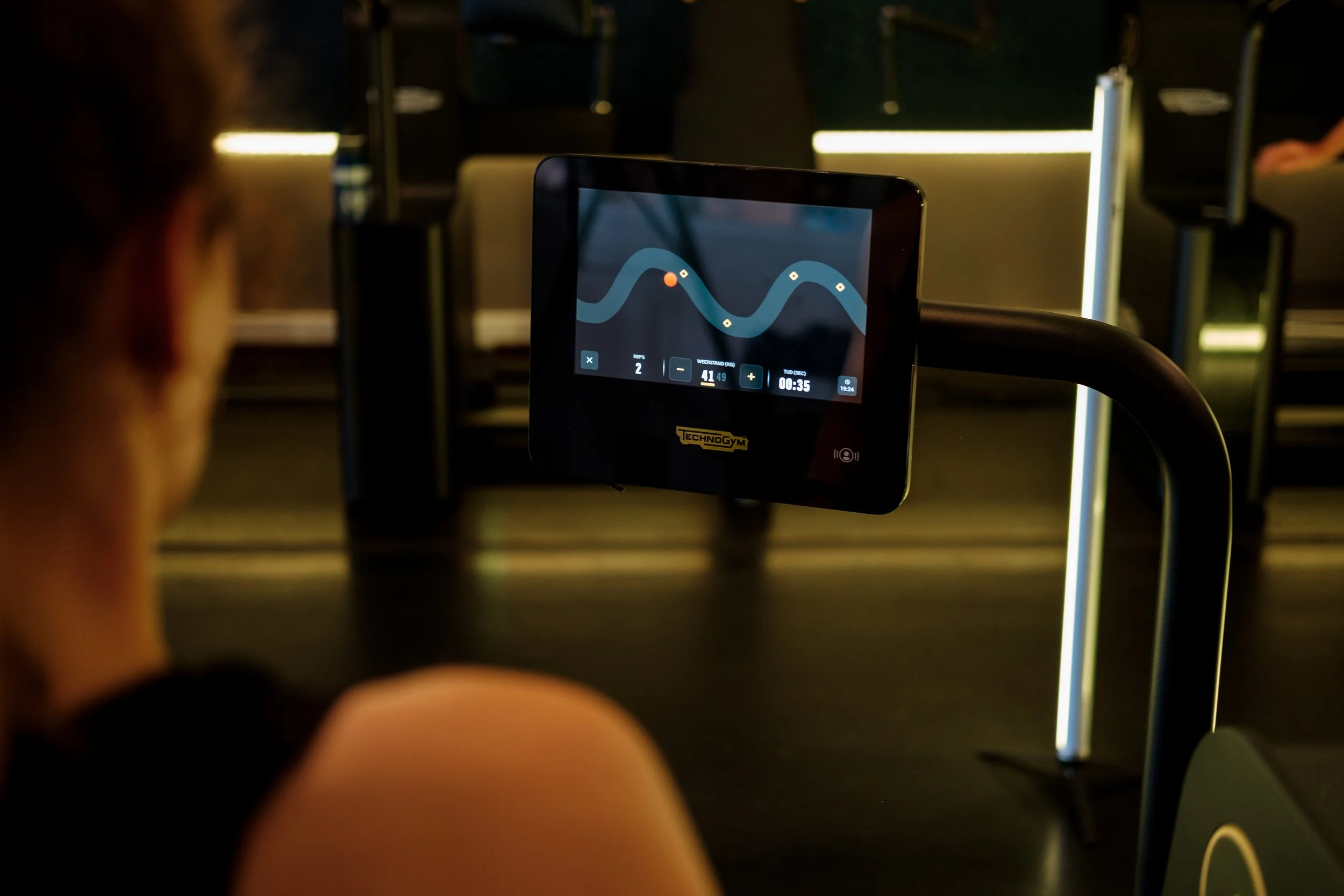 Een persoon gebruikt een fitnessapparaat met een digitaal scherm dat een golvende lijn en statistieken toont in een sportschool.