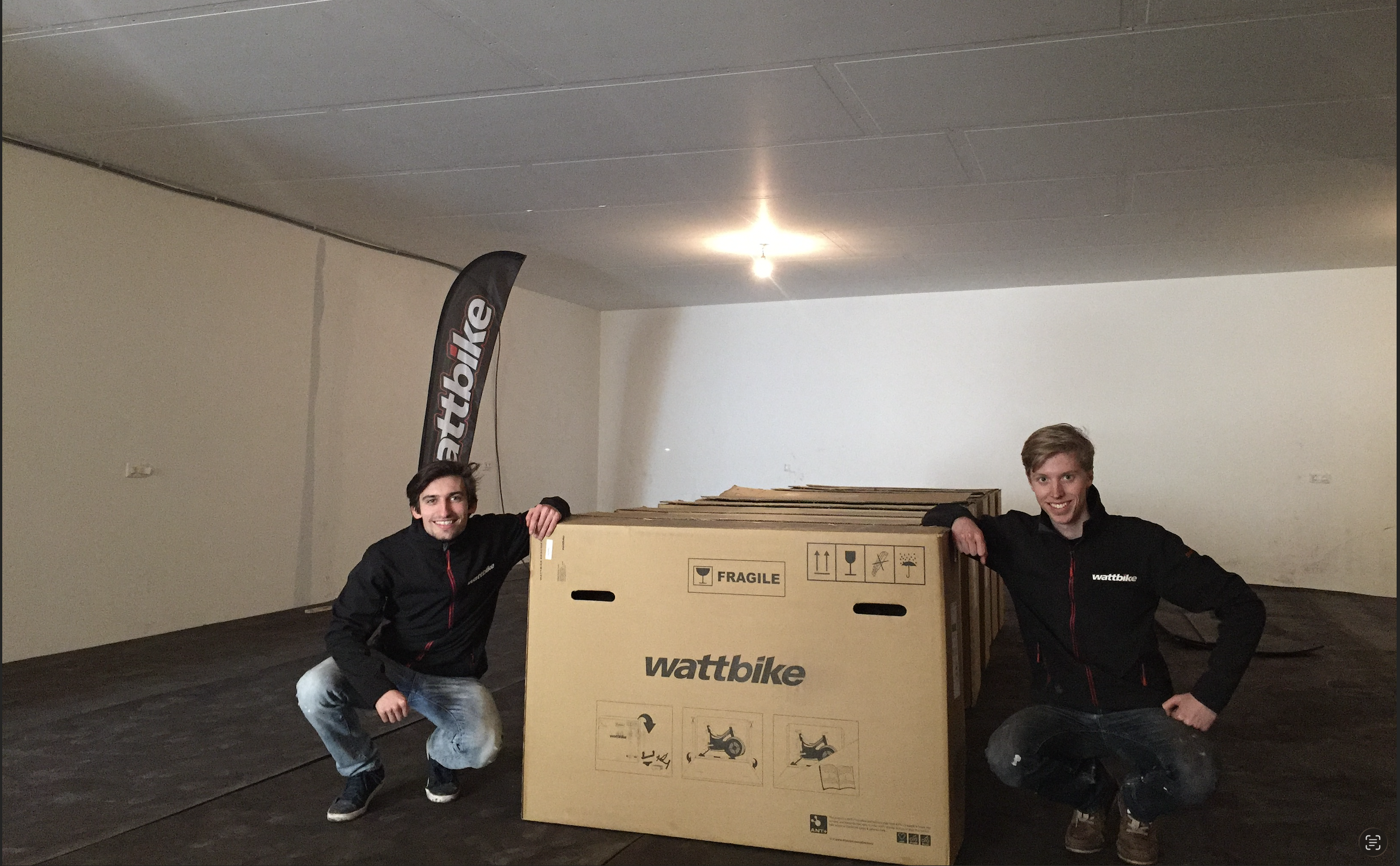 Twee mannen in zwart jack met rode accenten staan naast een grote kartonnen fietsdoos met het logo wattbike, in een lege kamer met witte muren.