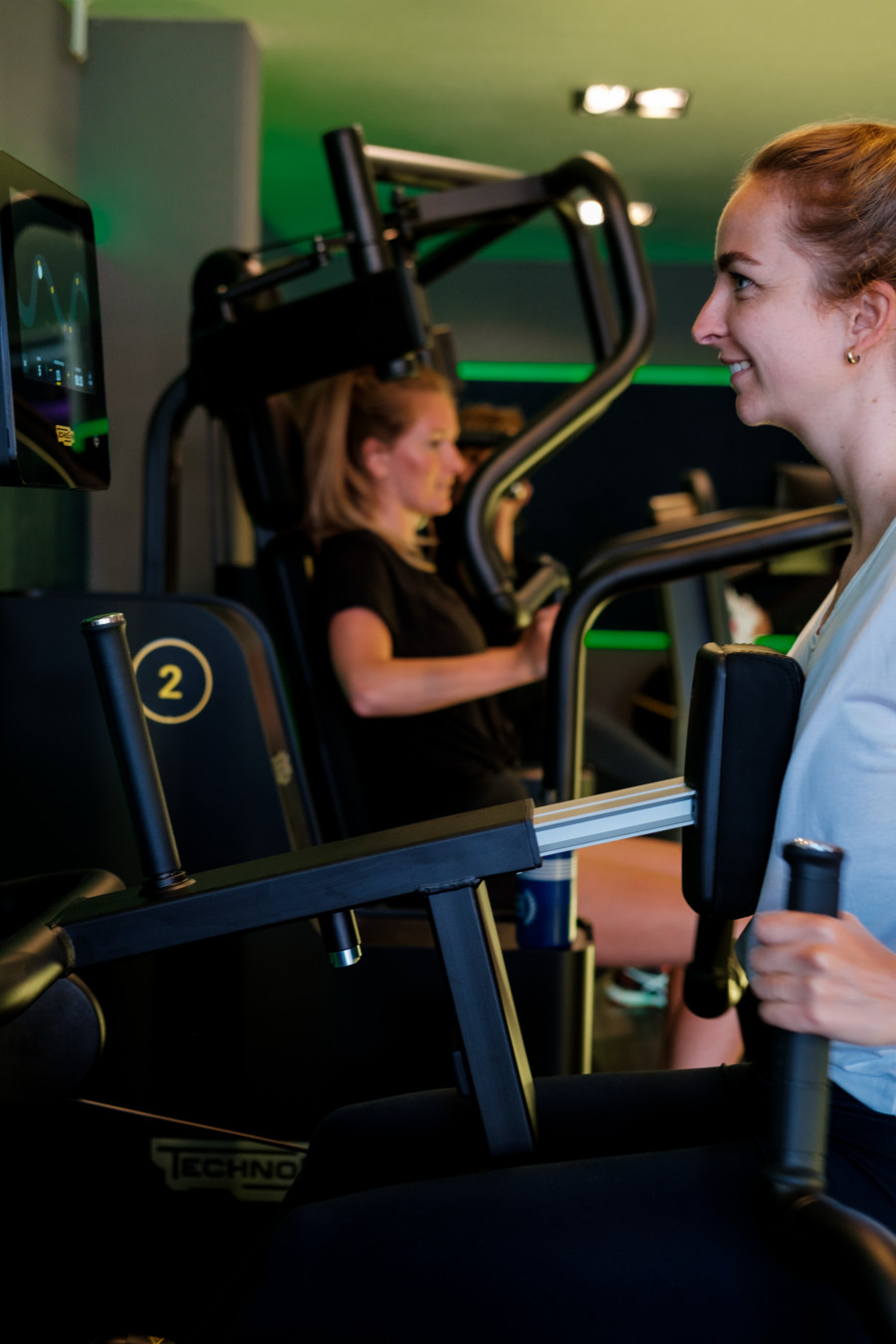 Twee vrouwen trainen op cardio-apparaten in een sportschool, met één vrouw voor de camera en een andere op de achtergrond.