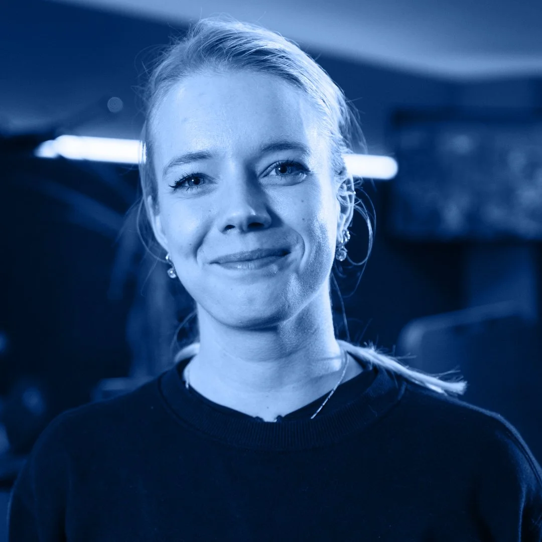 Portret van een lachende vrouw met blond haar, in een blauw getinte foto, in een donkere omgeving met lichtelementen op de achtergrond.