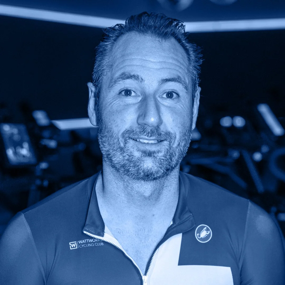 Een man met een baard en een glimlach, gekleed in een wielrenshirt, op een donkere achtergrond met fietsstations.