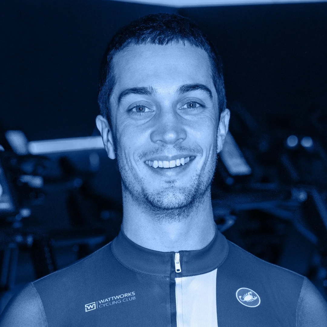 Portret van een lachende man in een wielerkleding met een logo van Wattworks Cycling Club, binnen een fitnessruimte of wielerwinkel.