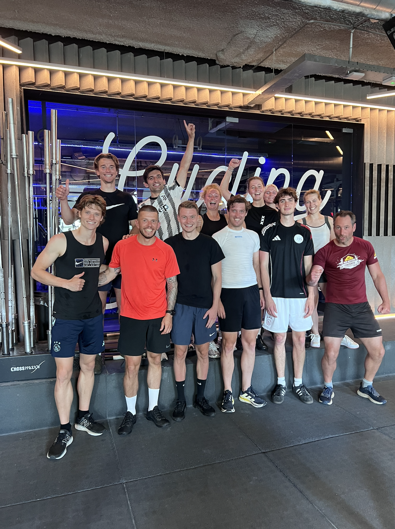 Gezelschap van elf mensen in sportkleding, blij en lachend, poseert binnen voor een winkelraam met de tekst 'Fysio'.