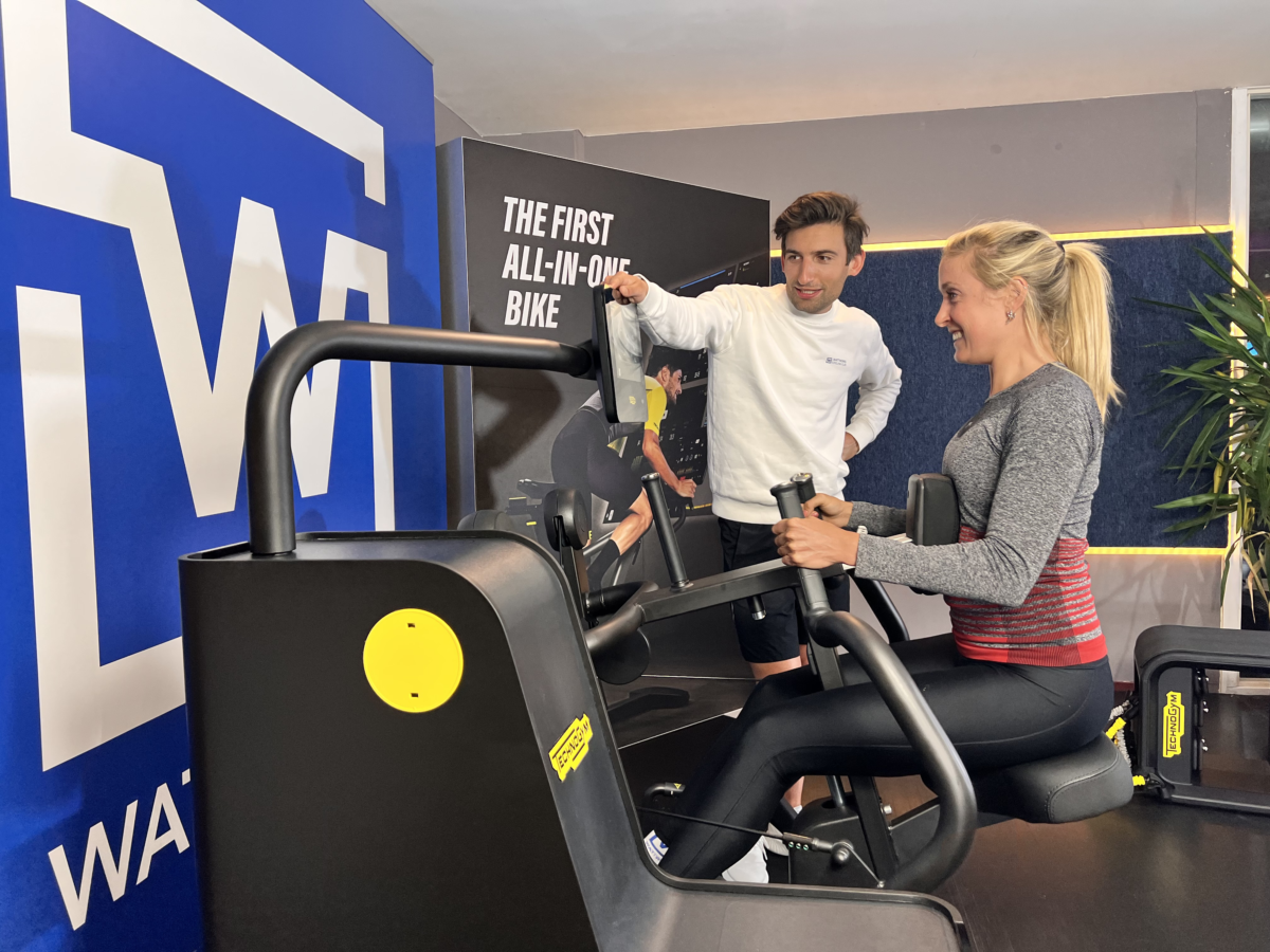 Een man en een vrouw in een sportschool, de vrouw zit op een fitnessapparaat en de man staat naast haar, beide lachen en lijken te praten.