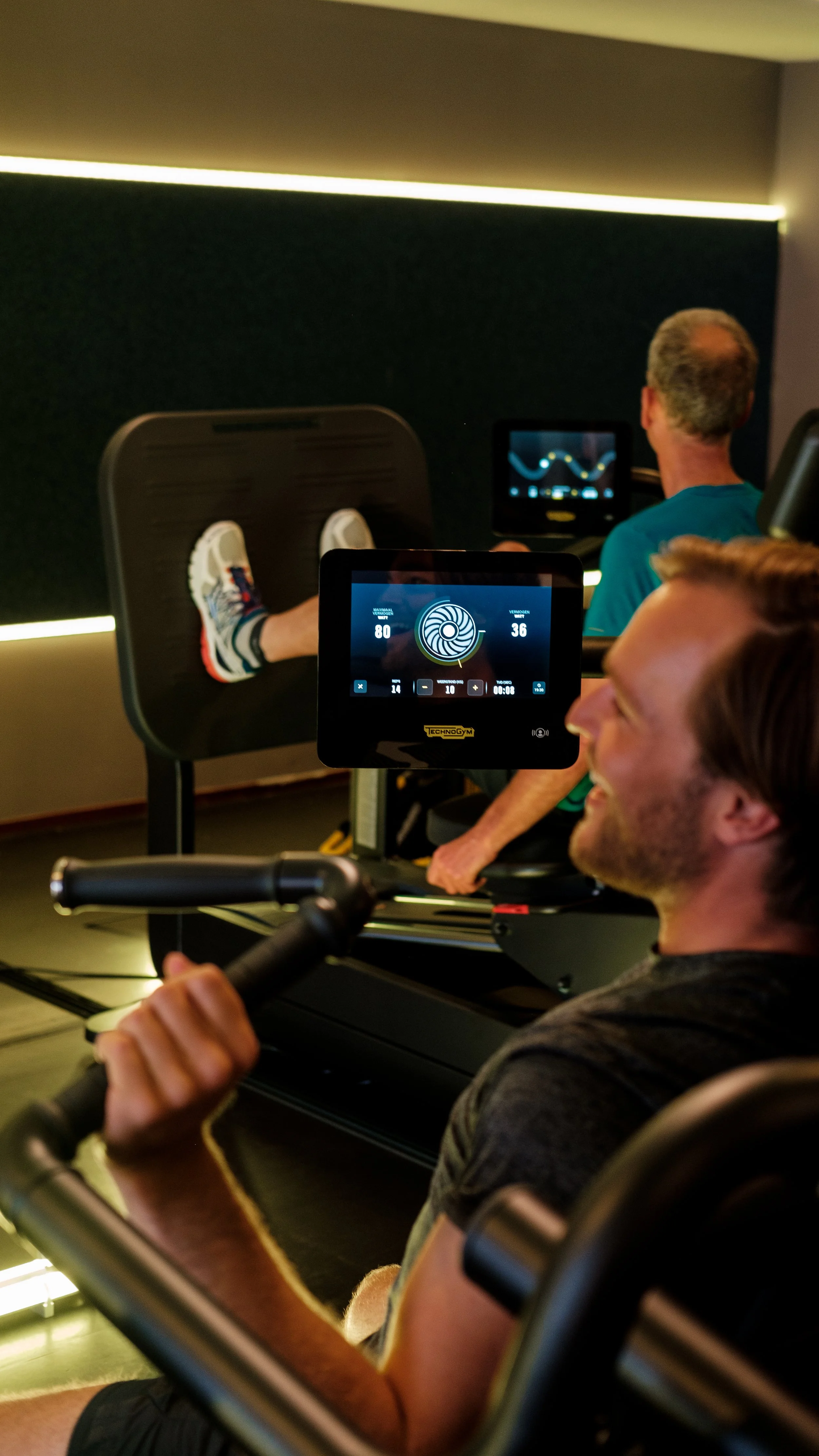 Mensen gebruiken een hometrainer in een fitnessruimte. Eén persoon zit op de hometrainer, anderen kijken naar een scherm met gegevens, terwijl een persoon op een stoel zit.