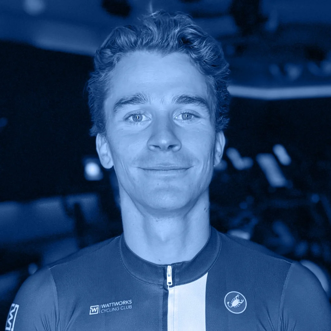 Een man met kort, bruin haar die een sportshirt draagt met het logo 'Wattwocks Cycling Club' op de linkerborst, lachend voor de camera. De afbeelding heeft een blauwe tint.