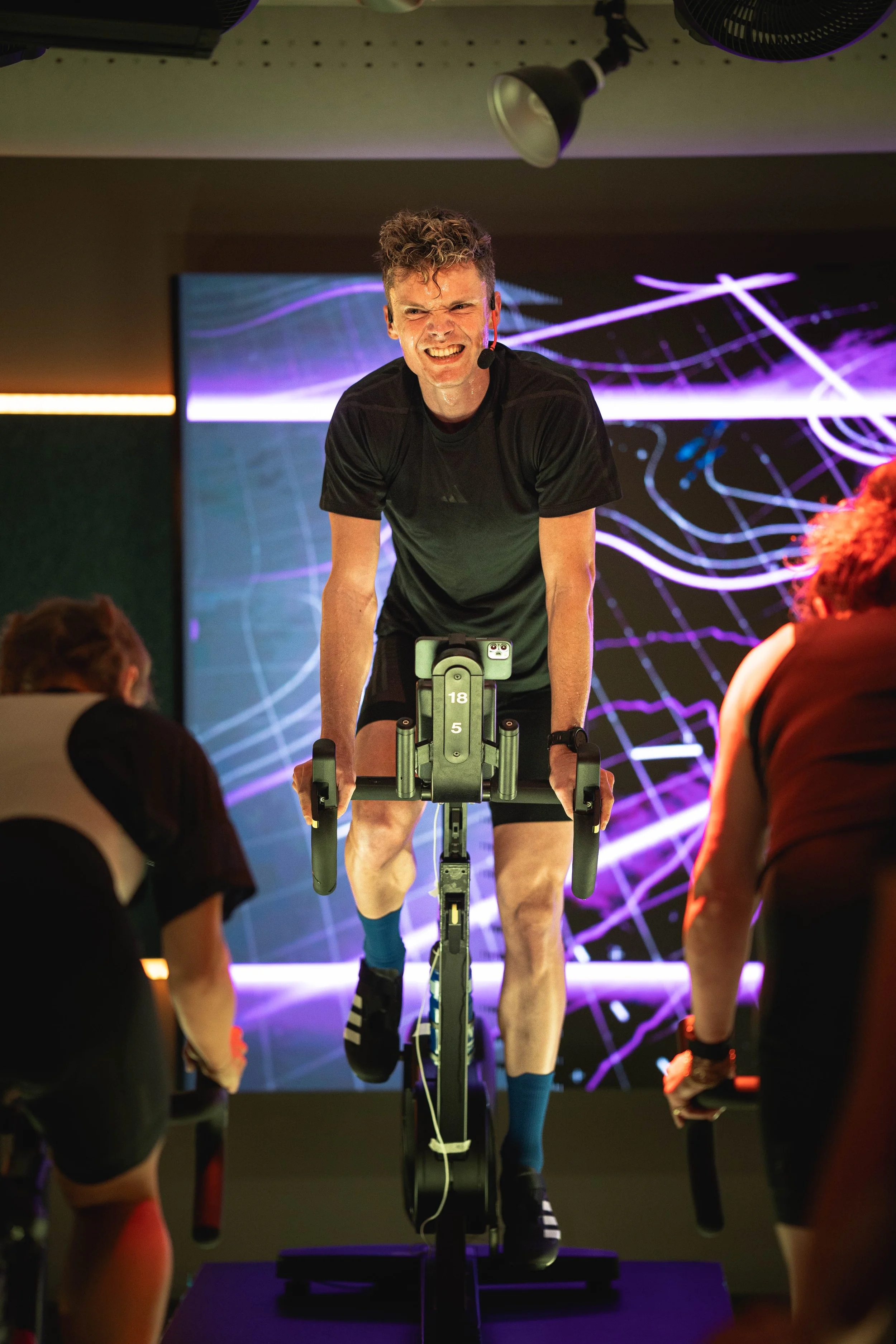 Man fietst op een binnentrainingsfiets, omringd door drie mensen die hem ondersteunen. De achtergrond heeft een kleurrijk, abstract neon-licht design.