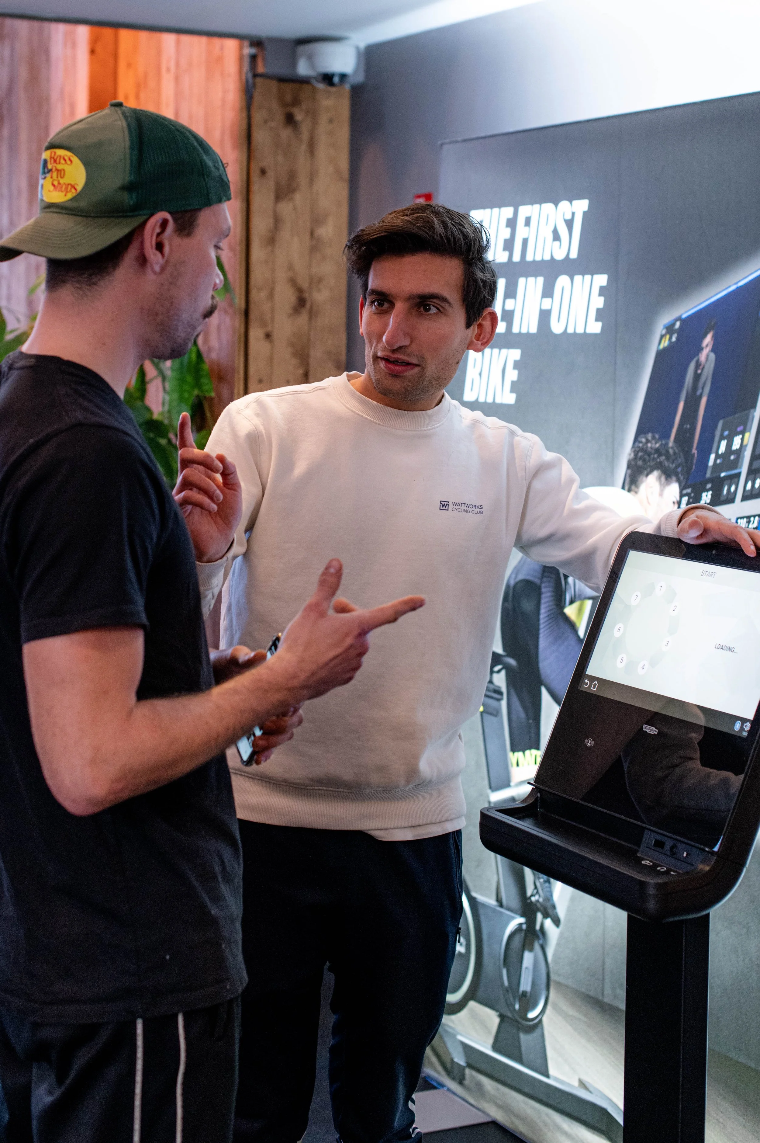 Twee mannen praten naast een digitaal kiosk met een fitness app op het scherm, een gym- of fitnessruimte op de achtergrond.