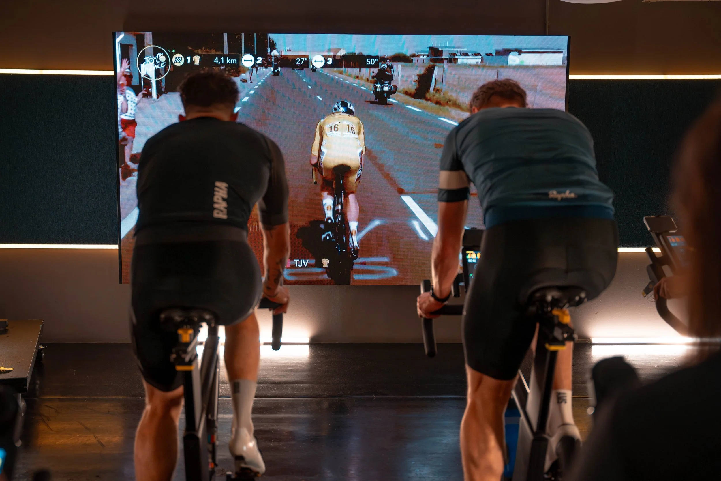 Mensen die op hometrainers fietsen tijdens een indoor fietstraining, met een grote scherm die een virtuele race toont.