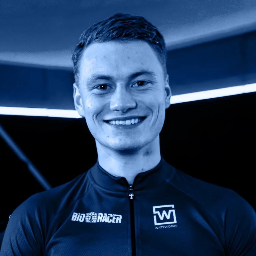 Portret van een jonge man met kort bruin haar, lachend, in een sportpak met logo's, binnen met donkere achtergrond en blauwe tinten.