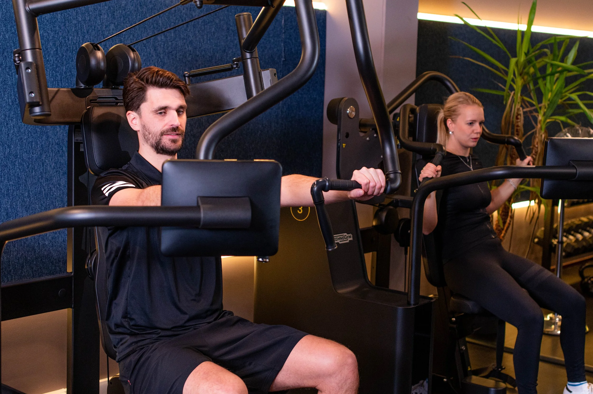 Een man en een vrouw die een fitnessapparaat gebruiken in een sportschool, omringd door planten en blauwe muur.