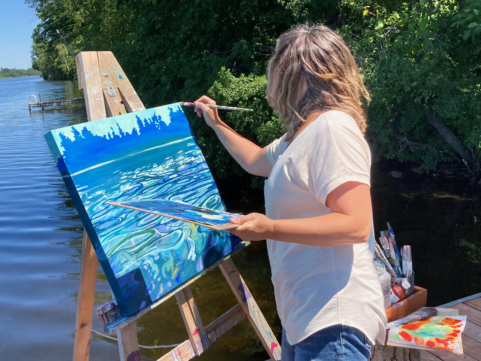 Mary painting a landscape of water, trees, and sky on an outdoor canvas near a lake, with paintbrushes and palette.
