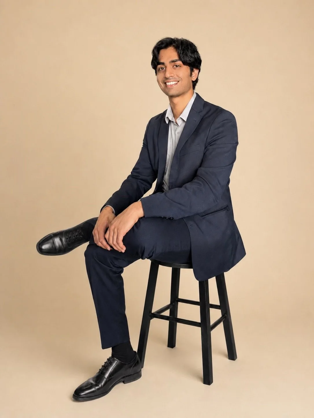 A man in a dark suit sitting on a black stool against a beige background.