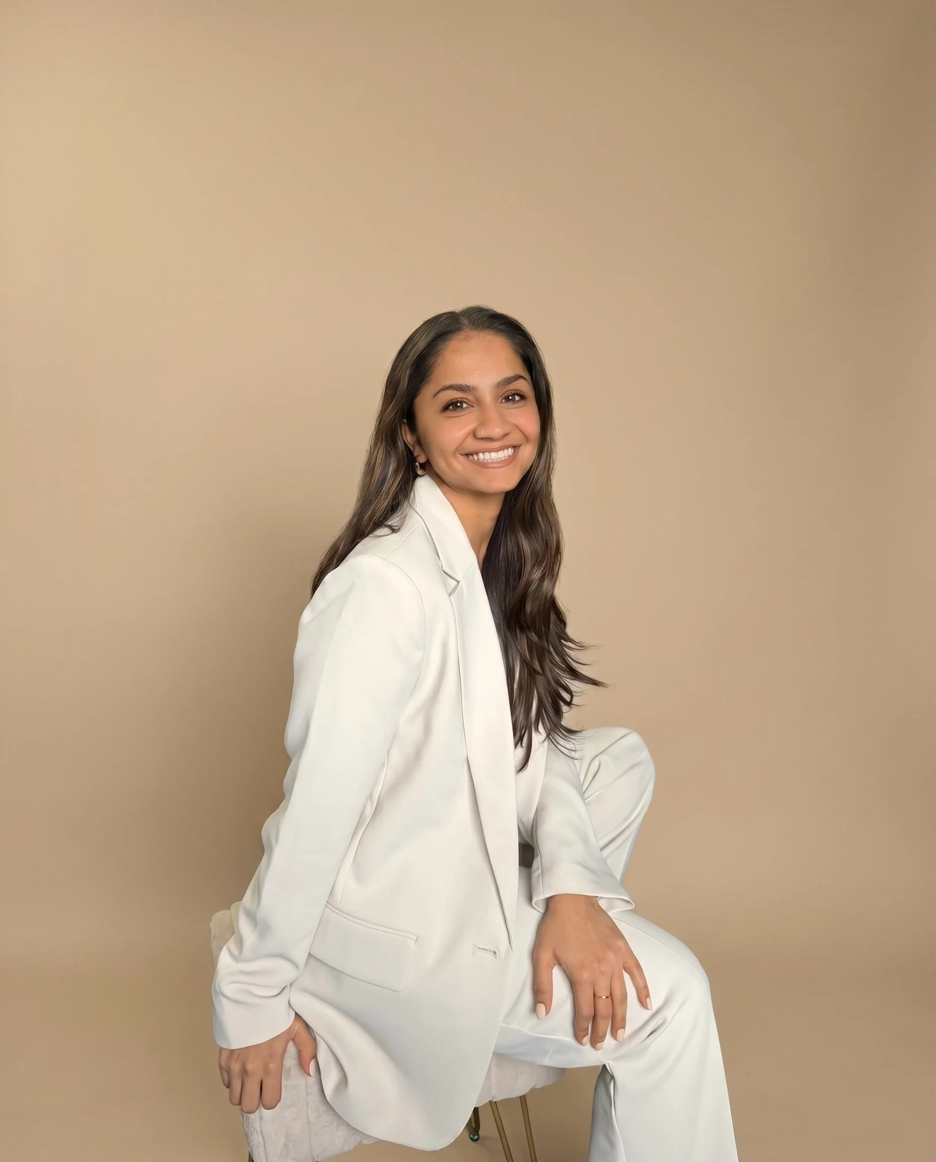 A woman with long brown hair smiling while seated in a white outfit against a beige background.