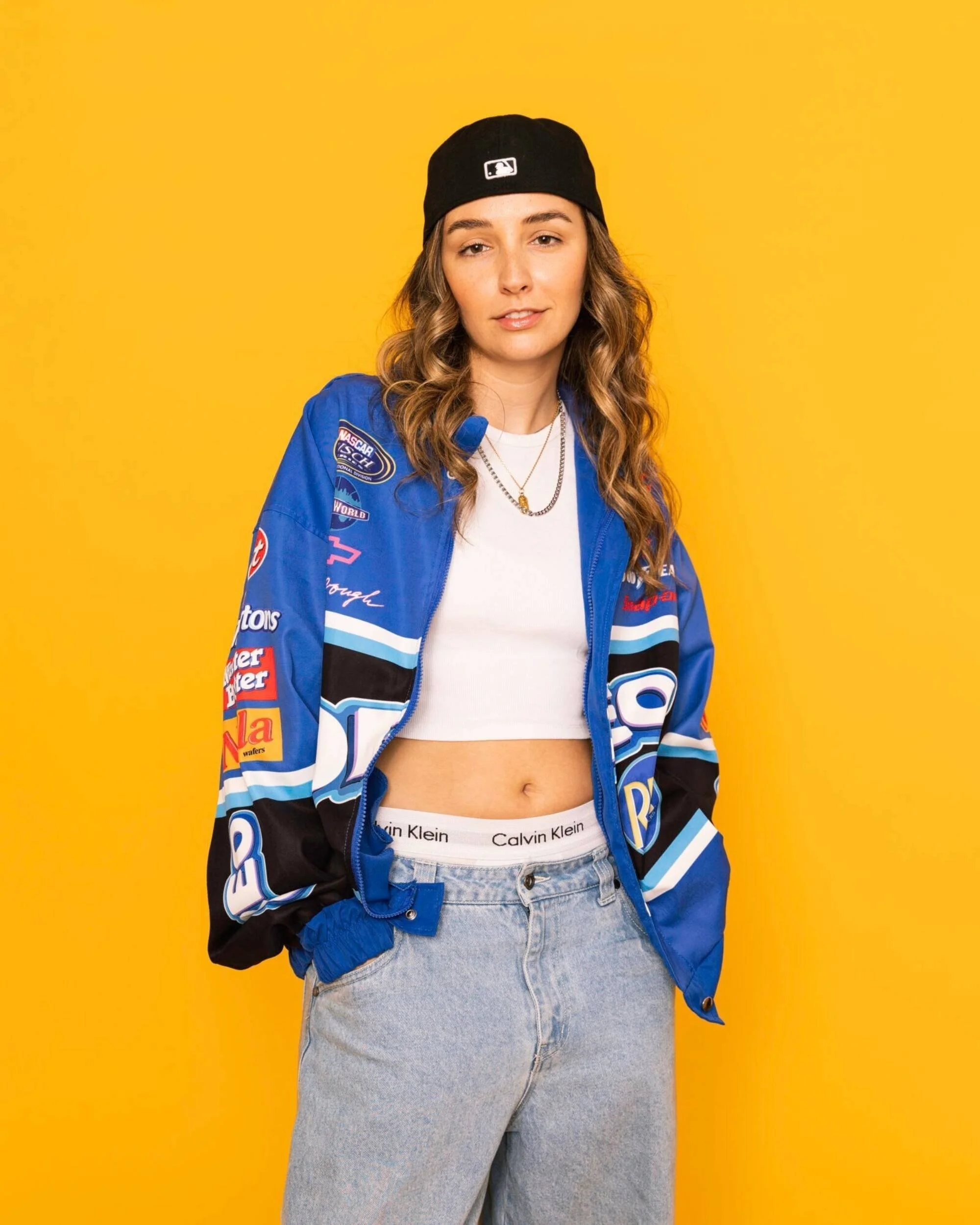 Jeune femme avec casquette noire, veste de course blue avec logos, crop top blanc, jean bleu, fond jaune