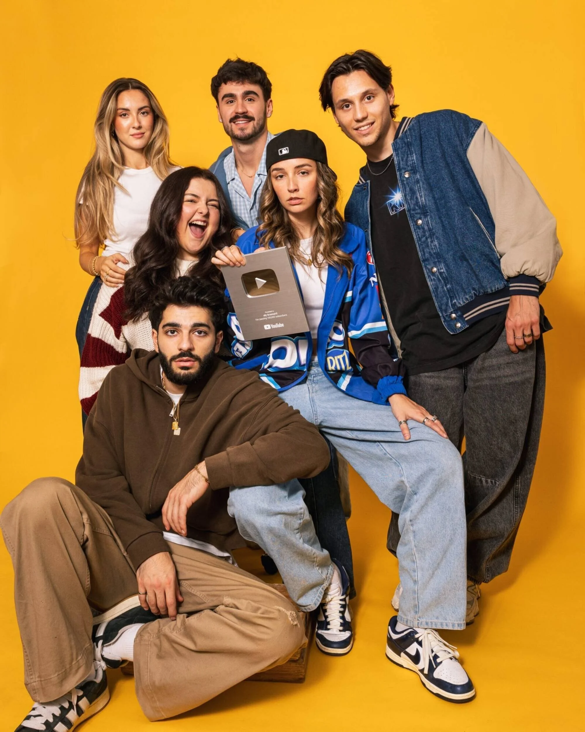 Groupe de six jeunes adultes posant devant un fond jaune. L'un d'eux tient une plaque de reconnaissance YouTube.