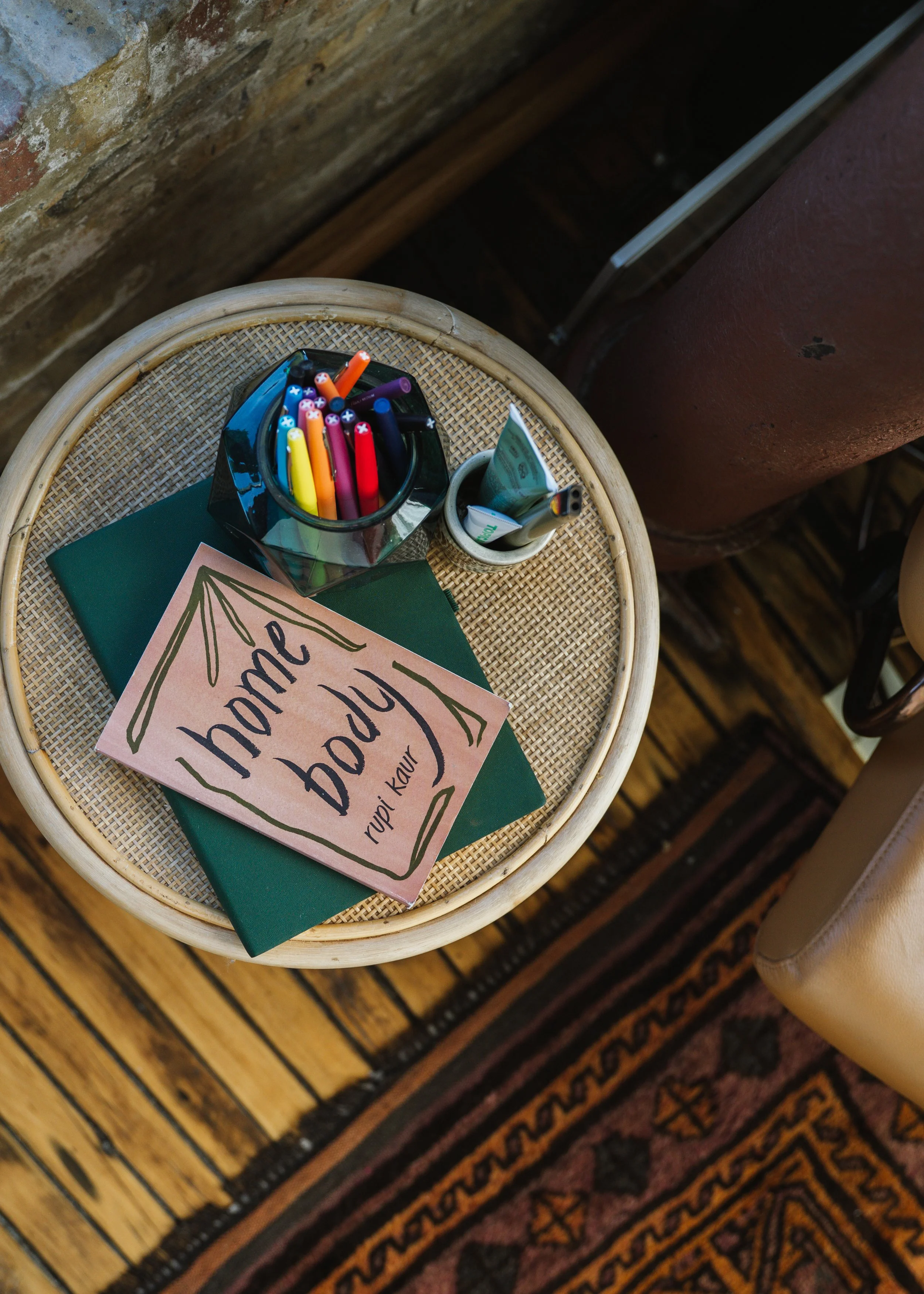 A round wicker table holding a dark glass container with colorful markers, a small white container with pens, a green book, and a pink card with black handwritten text that says 'home body' by Rupi Kaur. The table is placed on a wooden floor next to a brick wall with an exposed pipe.
