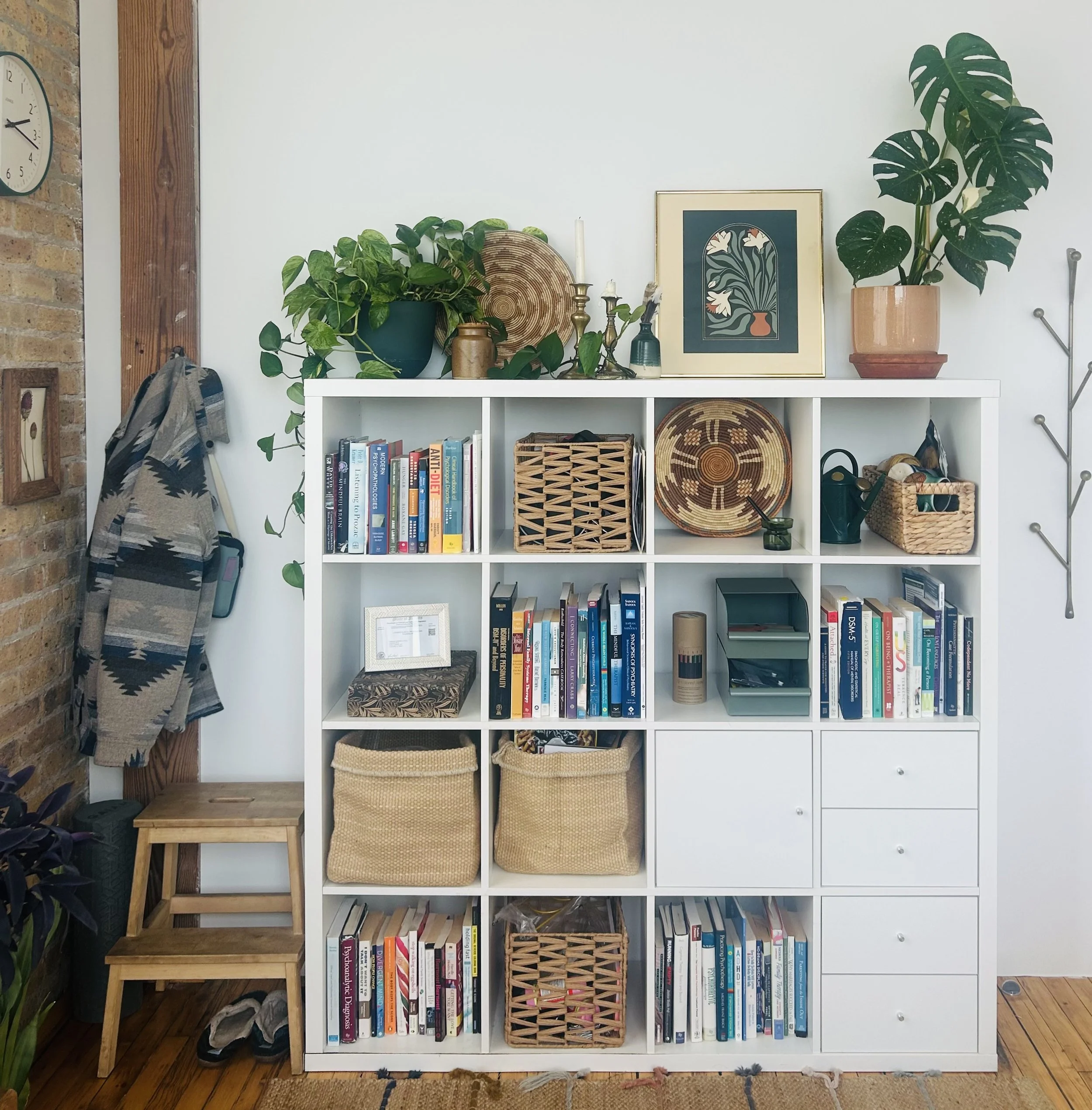 White bookshelf with books, baskets, and decorative items, topped with potted plants and artwork, in a cozy living space.