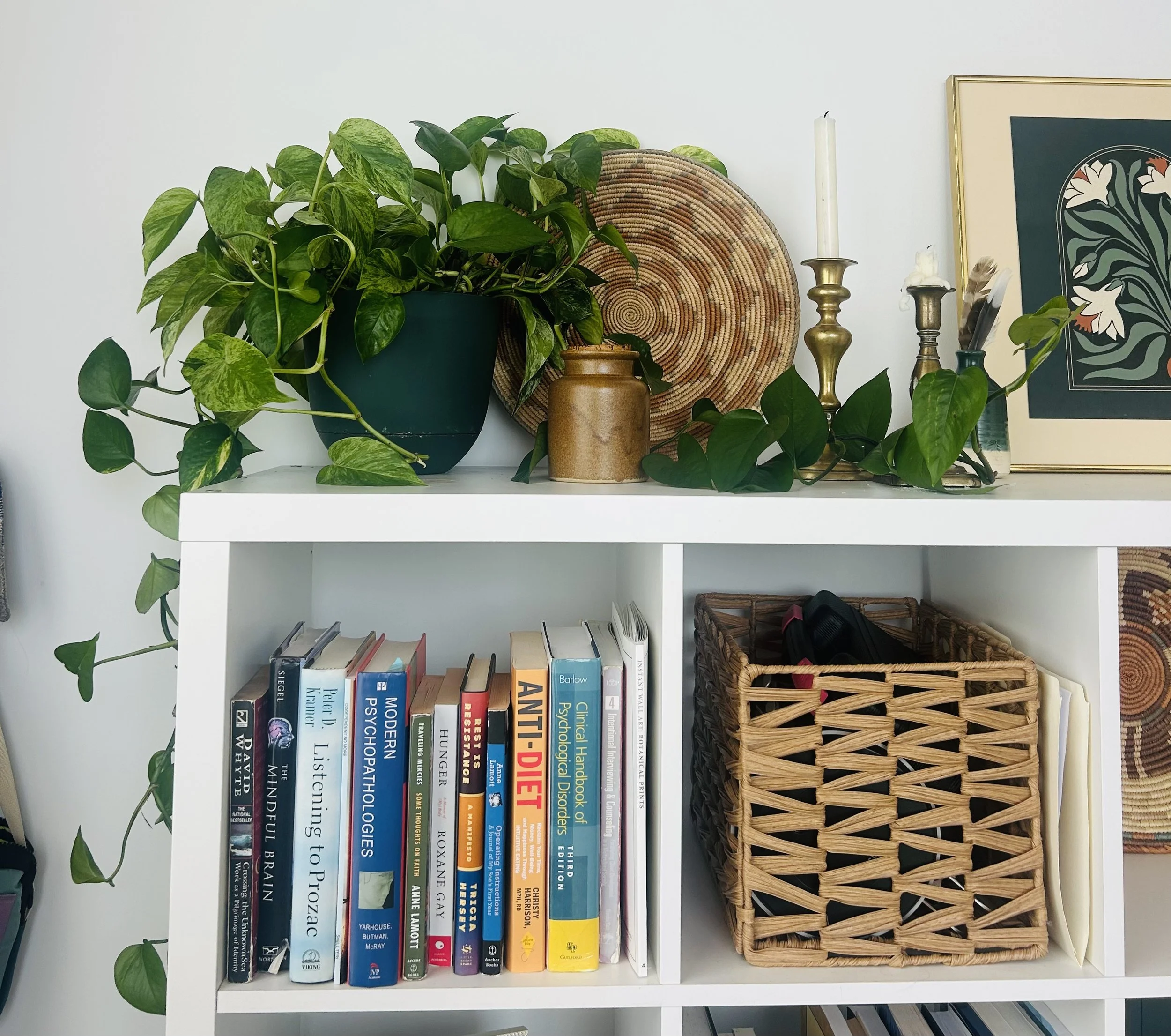 White bookshelf with books, a wicker basket, a plant in a dark green pot, and decorative objects including a framed artwork and candlesticks.