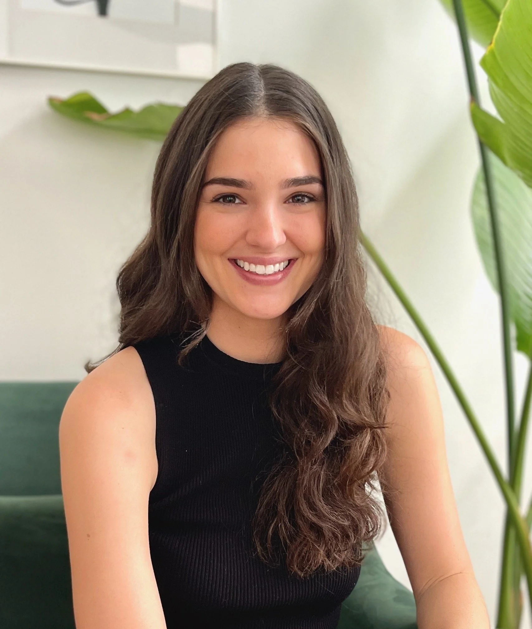 A young woman with long, wavy brown hair smiling, wearing a black sleeveless top, sitting indoors near green plants.