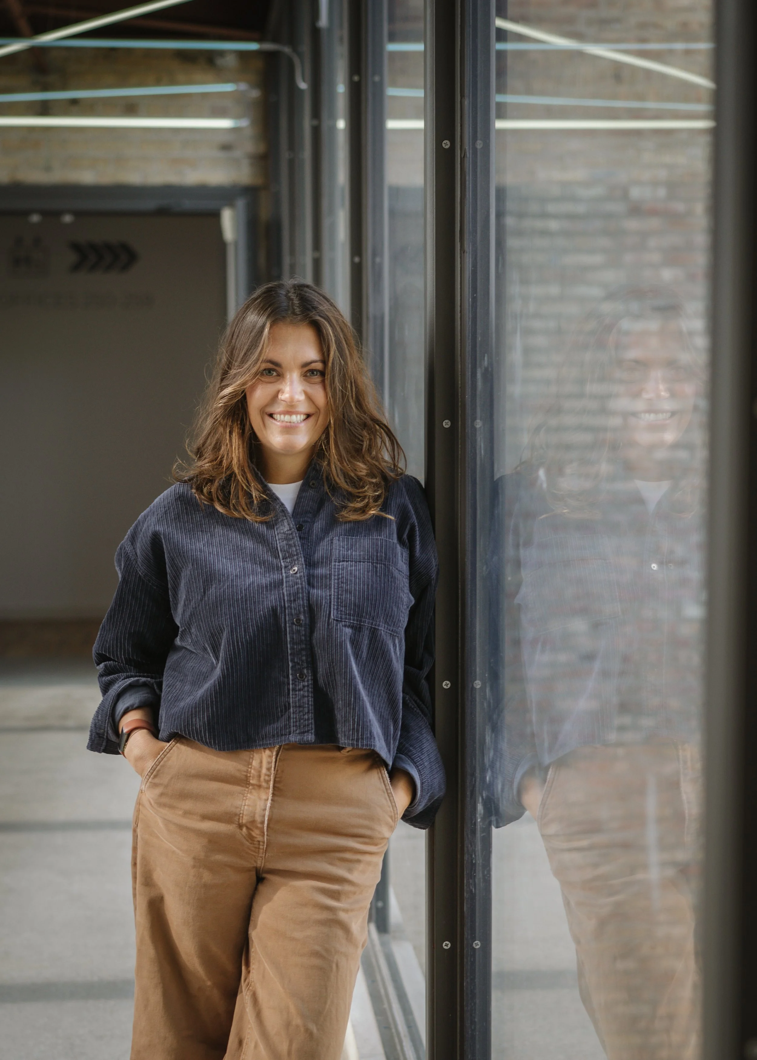 A smiling woman standing beside a glass wall in an industrial setting, with her hands in her pockets, wearing a dark blue button-up shirt and tan pants.