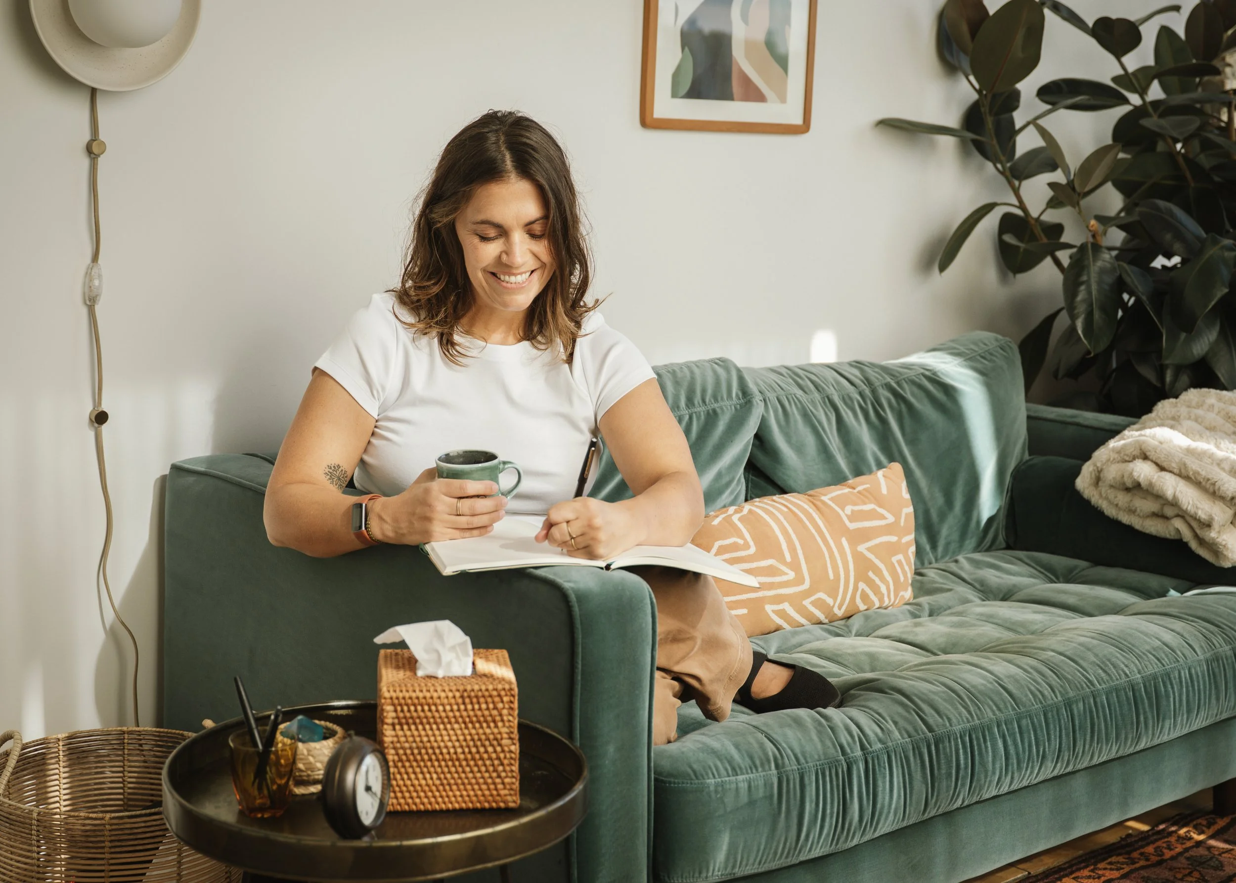 A woman sitting on a green velvet sofa, smiling as she writes in a notebook and holds a mug, in a cozy living room.