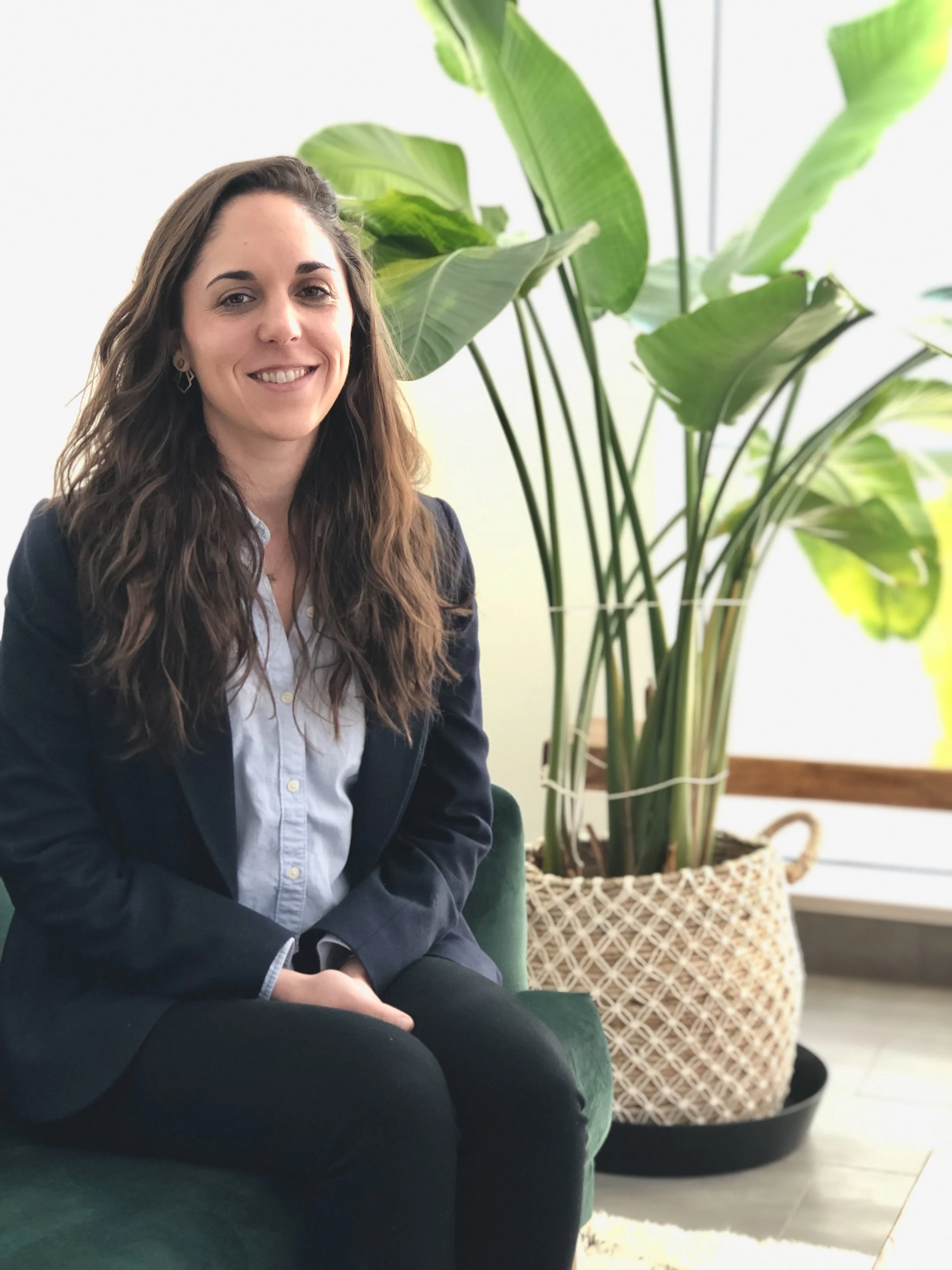 A woman with long brown hair, wearing a white shirt and a dark blazer, sitting on a green chair next to a large potted plant with broad green leaves.