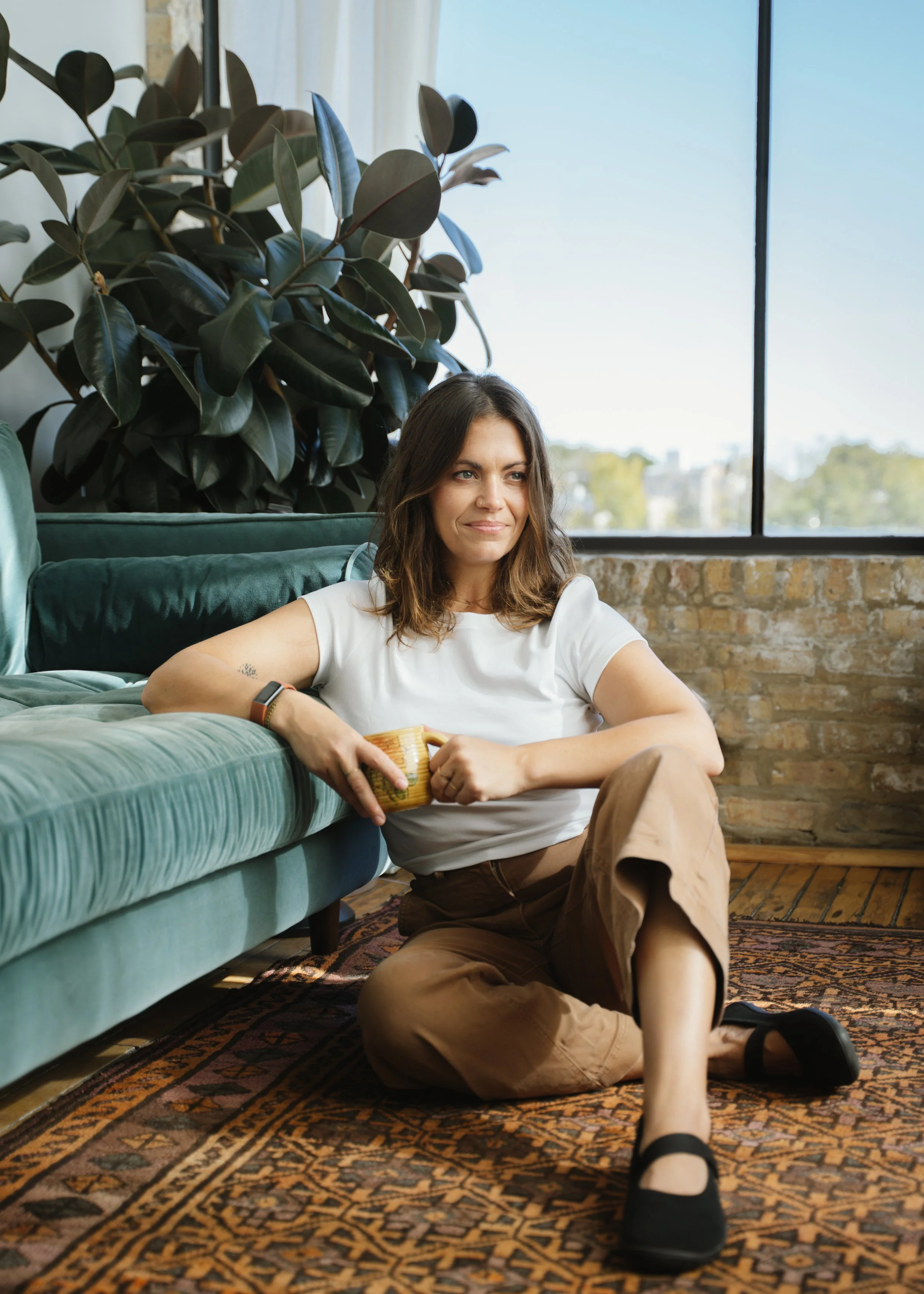 Woman sitting on the floor next to a green velvet couch, holding a coffee mug, with a large houseplant and window in the background.