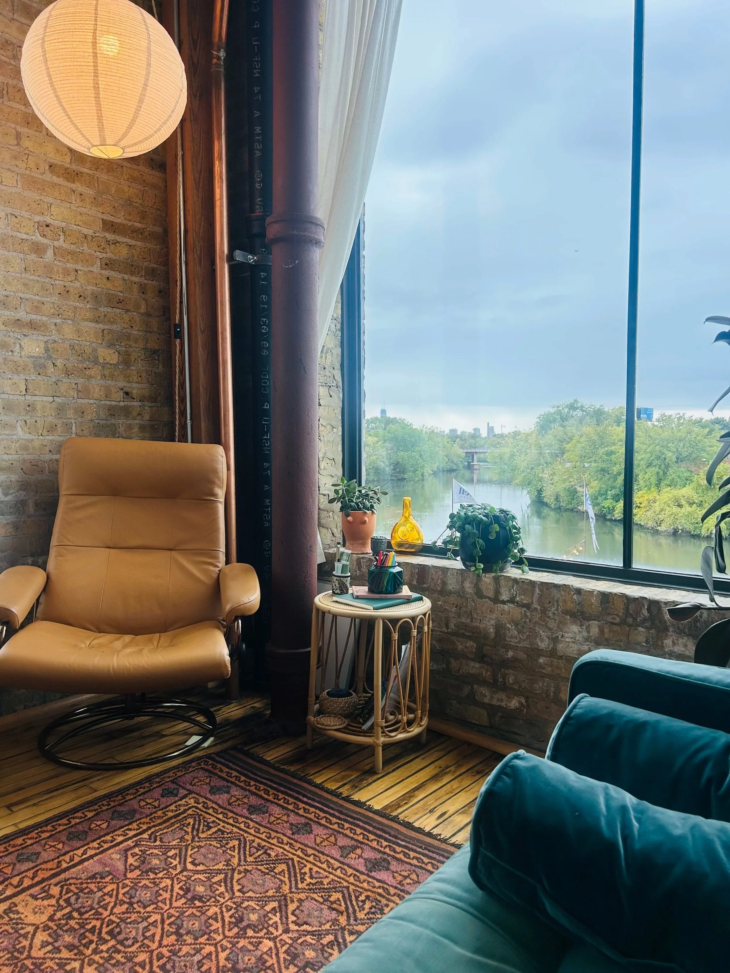 Cozy living room corner featuring a tan leather armchair, a small round wicker side table with plants, snacks, and writing utensils, a large window with a view of a river and greenery, exposed brick walls, a colorful patterned rug, and a large potted plant.
