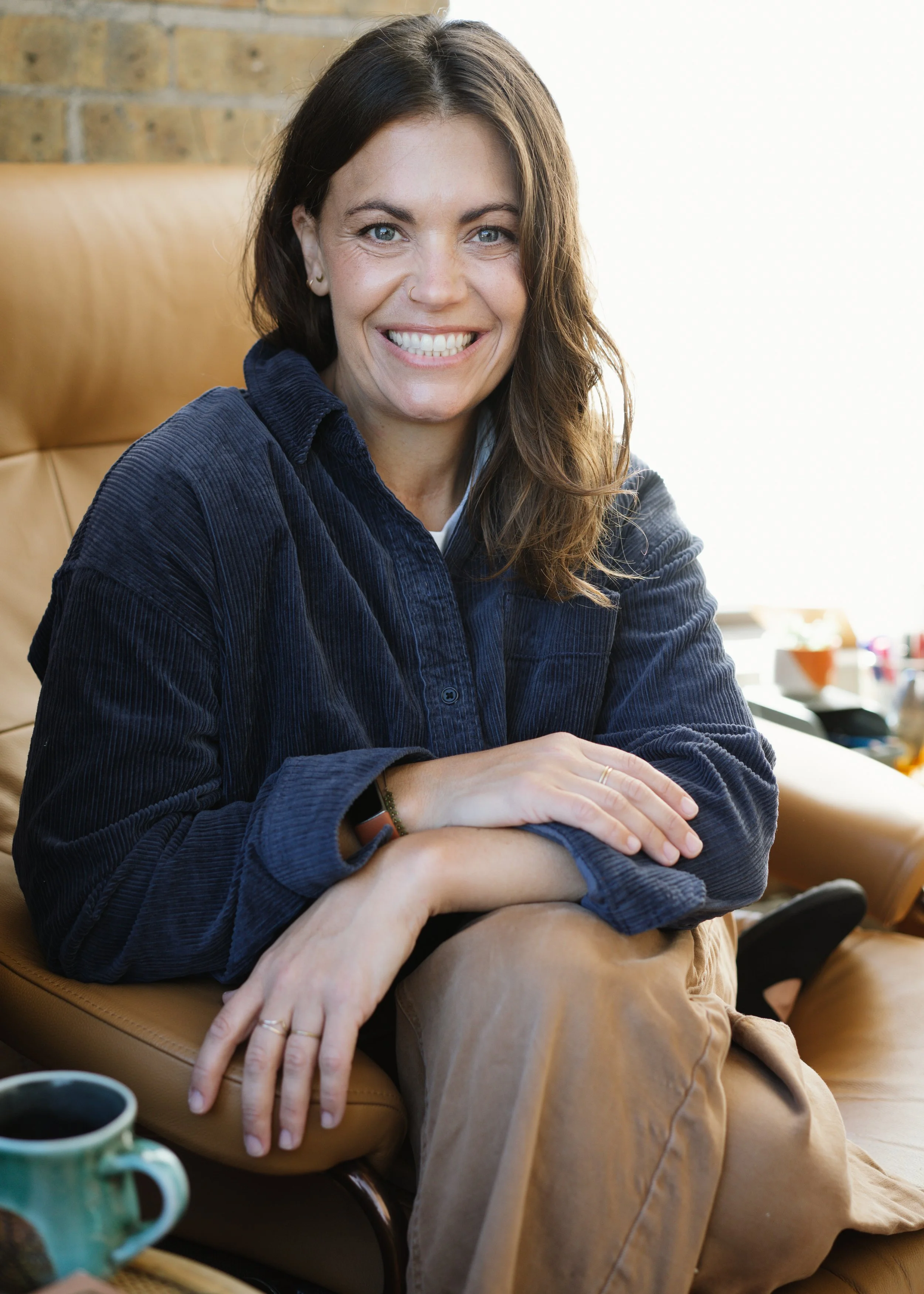 A woman with brown hair and blue eyes sitting on a tan leather chair, smiling with arms crossed, wearing a navy blue shirt and tan pants, in a cozy indoor setting with a brick wall and window in the background.