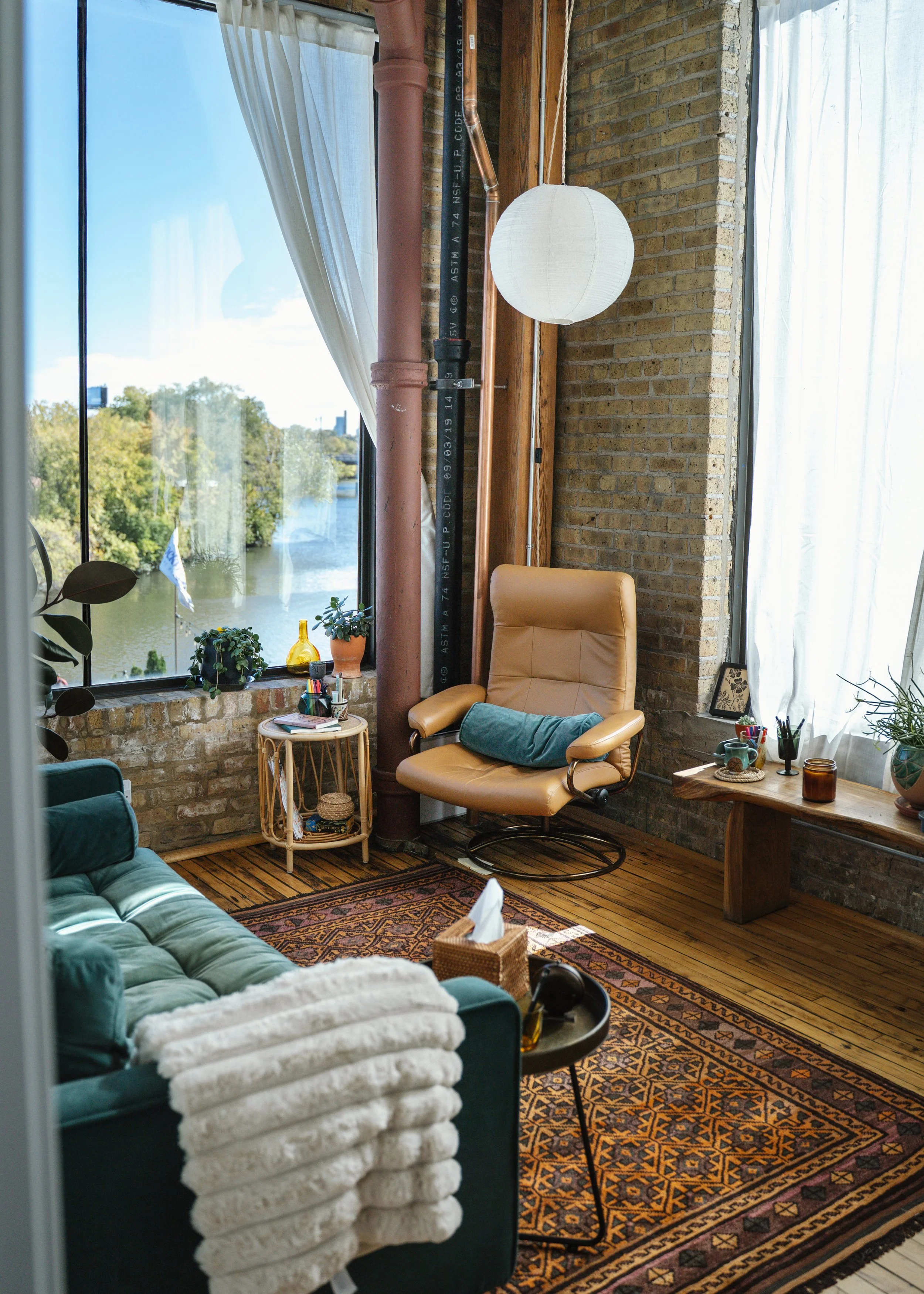 Cozy living room with large window overlooking a river, featuring a teal velvet sofa with white throw blanket, a tan leather accent chair, a wooden table with plants and art supplies, a small side table, a patterned rug, and white curtains.
