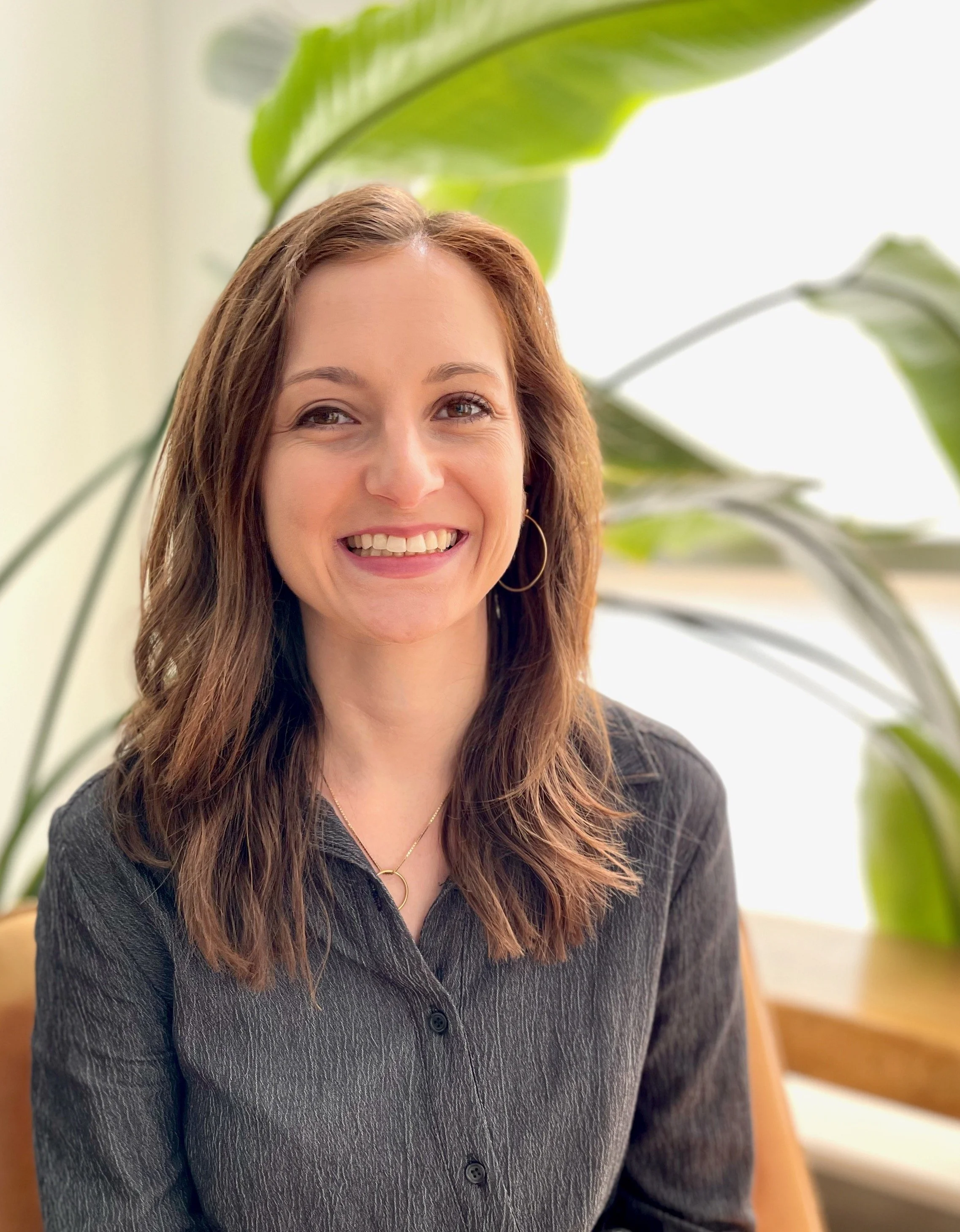 A woman with shoulder-length brown hair smiling, wearing a dark gray shirt, gold hoop earrings, and a gold necklace, seated near large green tropical plants.