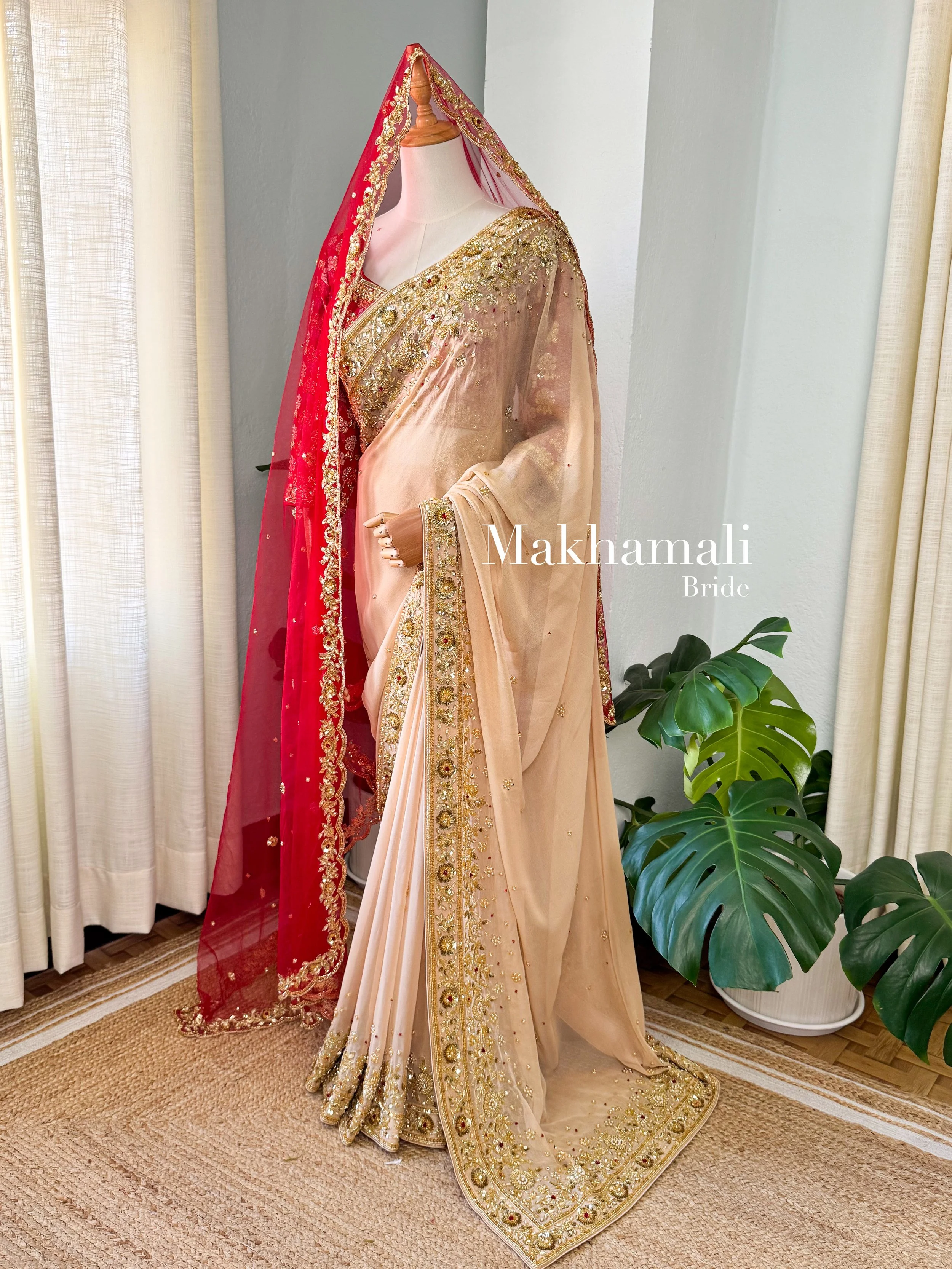 A beige and gold embroidered saree with a matching dupatta, displayed on a mannequin for a wedding or special occasion, with a large green leafy plant nearby.