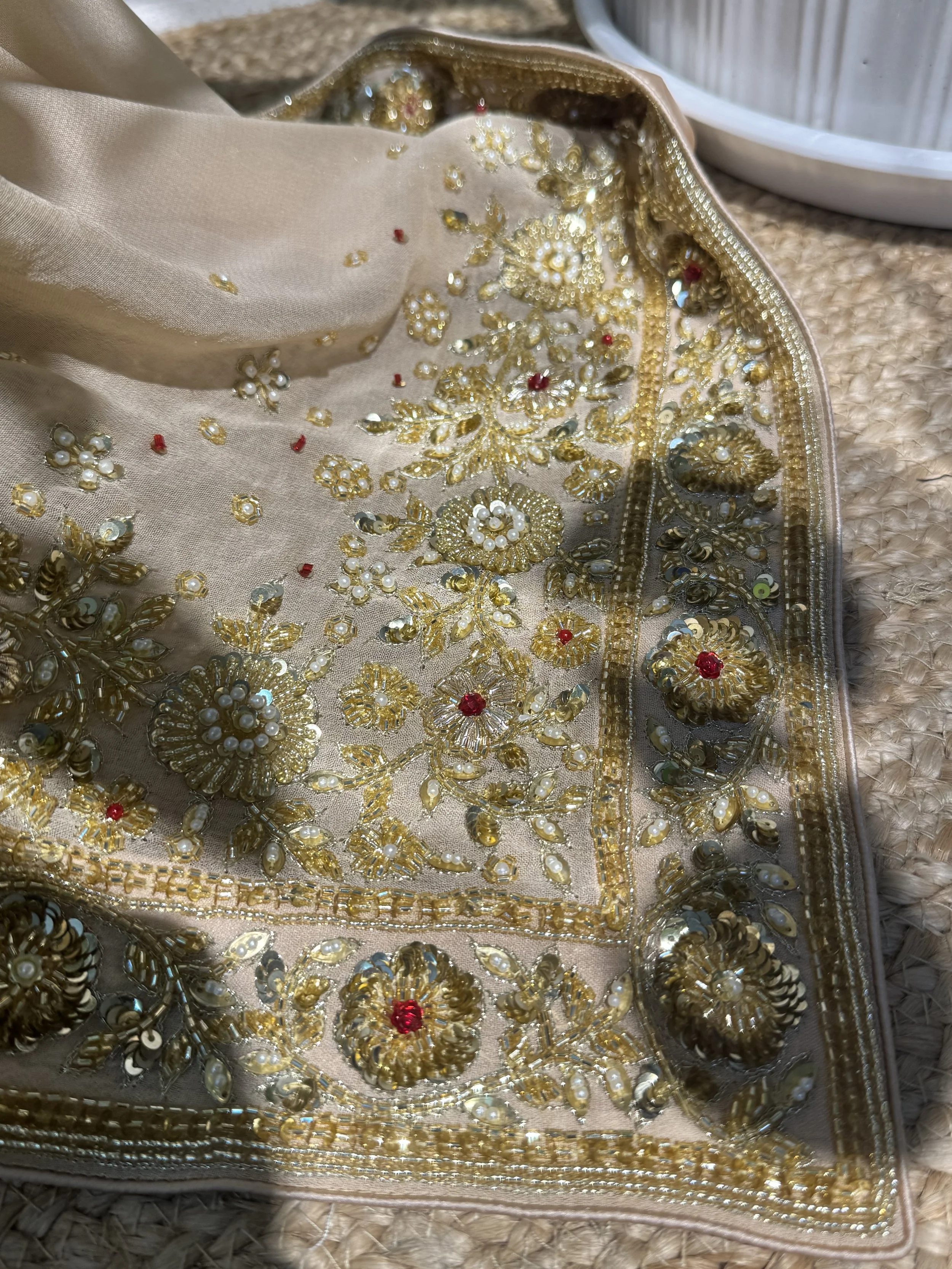 Close-up of beige fabric with intricate gold embroidery, pearls, and red gemstones, on a woven surface.