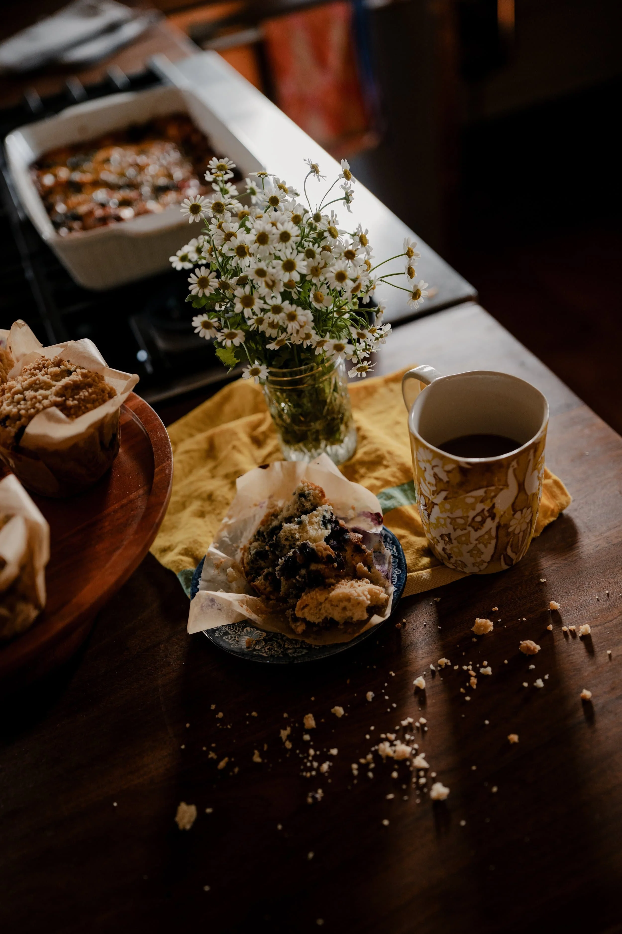 A vase of small white daisies with yellow centers on a wooden table with scattered cookie crumbs, a cup of coffee, and baked goods, with a baking dish in the background.